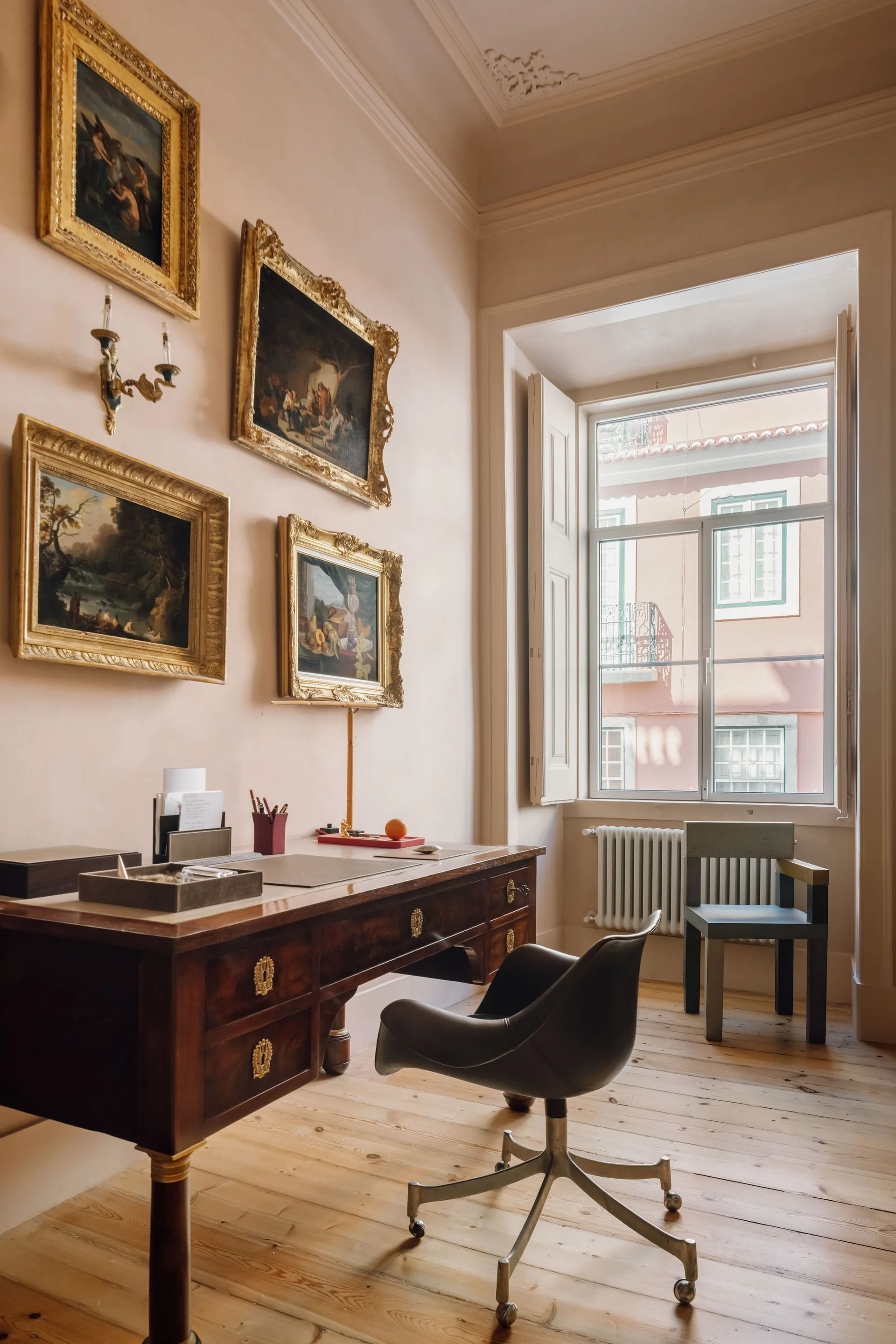A traditional dark wood writing desk sits on light pine floors in a pale pink study with gold-framed paintings and a window.