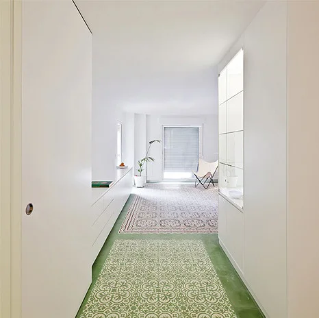 A minimalist white hallway features seamless built-in cabinetry and a green and white patterned tile floor leading to a bright room.