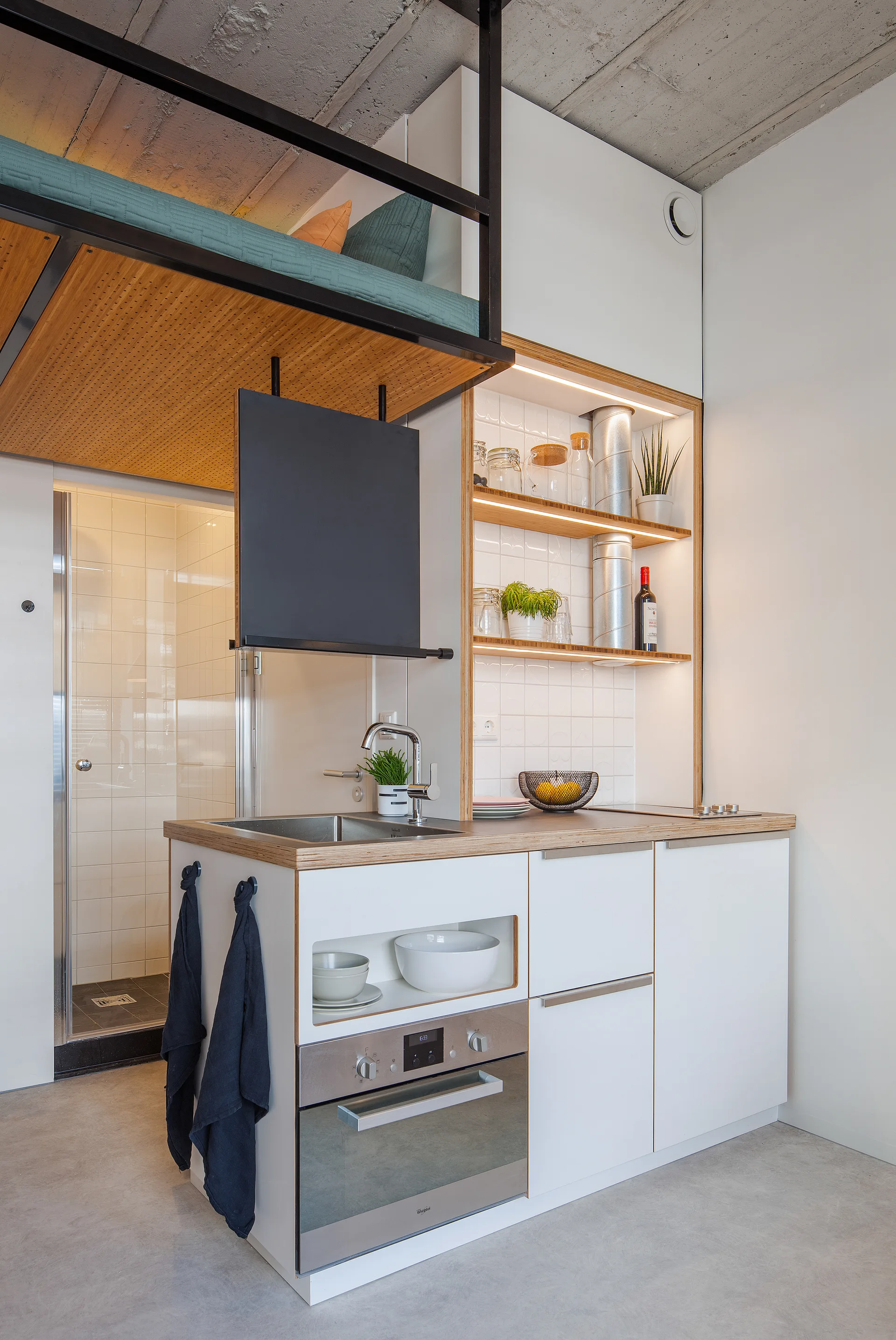 A compact white kitchenette with a wood-trimmed counter and shelves features a built-in oven and is situated beneath an overhead loft sleeping area.