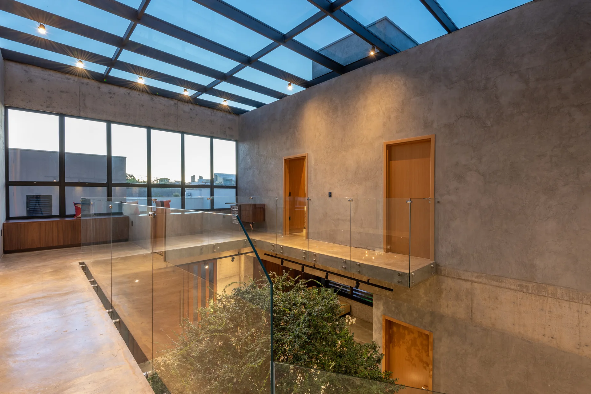 A concrete mezzanine landing with a glass railing stands beneath a large grid skylight, overlooking an indoor atrium with green plants.