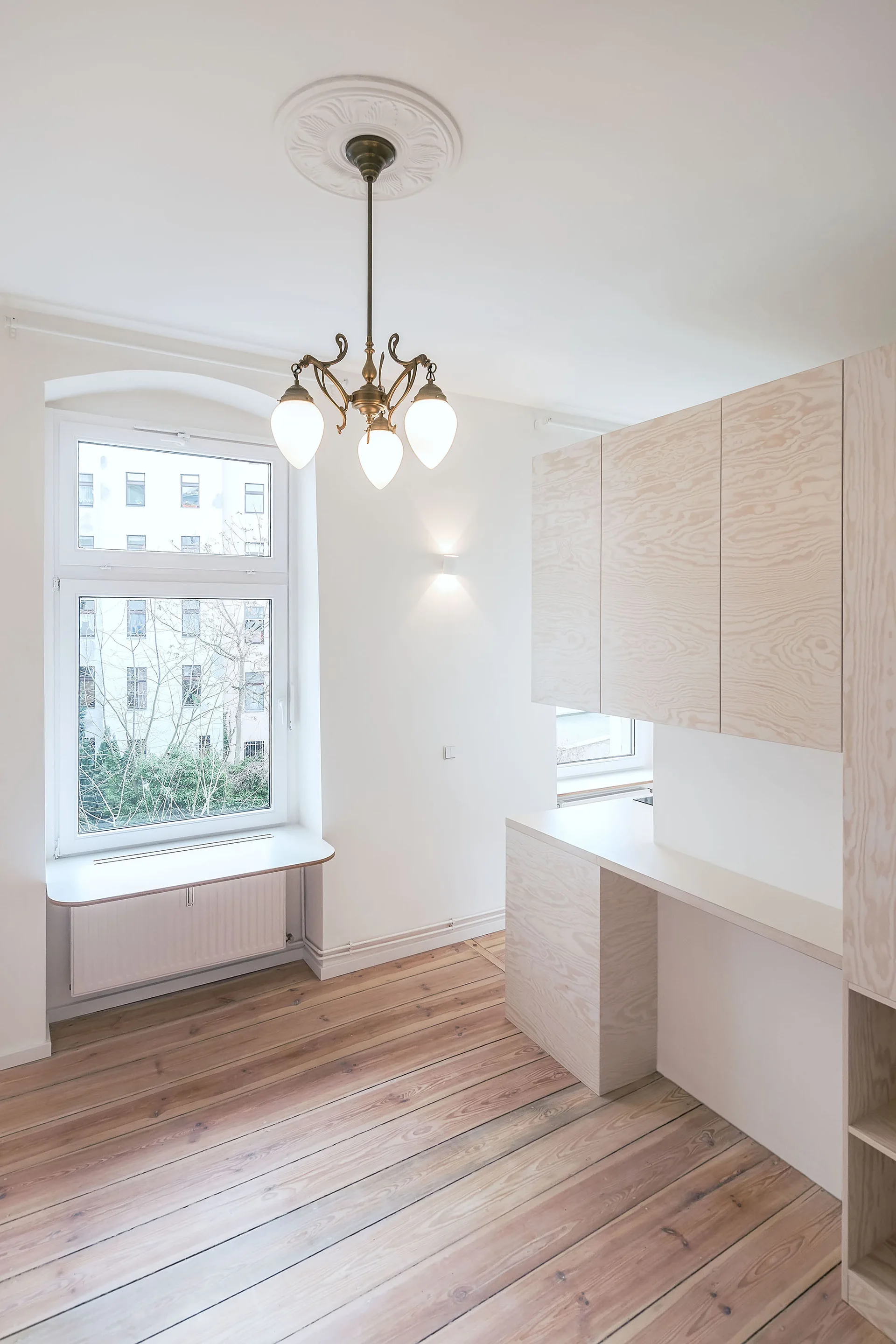 A bright room featuring modern pale plywood cabinetry, wide plank wood floors, and a traditional brass chandelier hanging from a ceiling medallion.