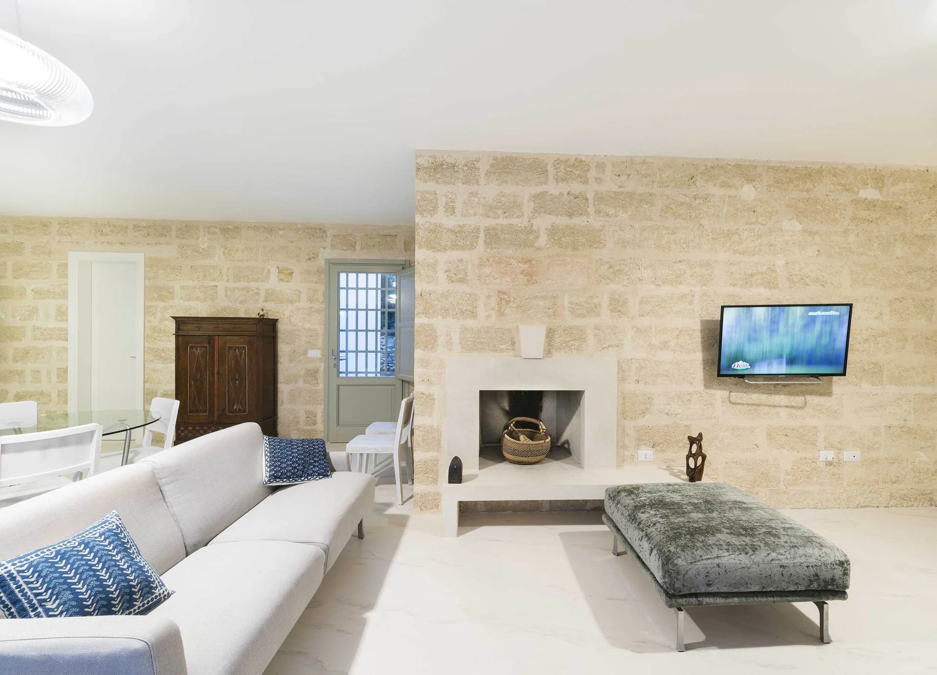 A contemporary grey sofa and a grey velvet ottoman sit on light tiled floors in a living room with beige exposed stone block walls.