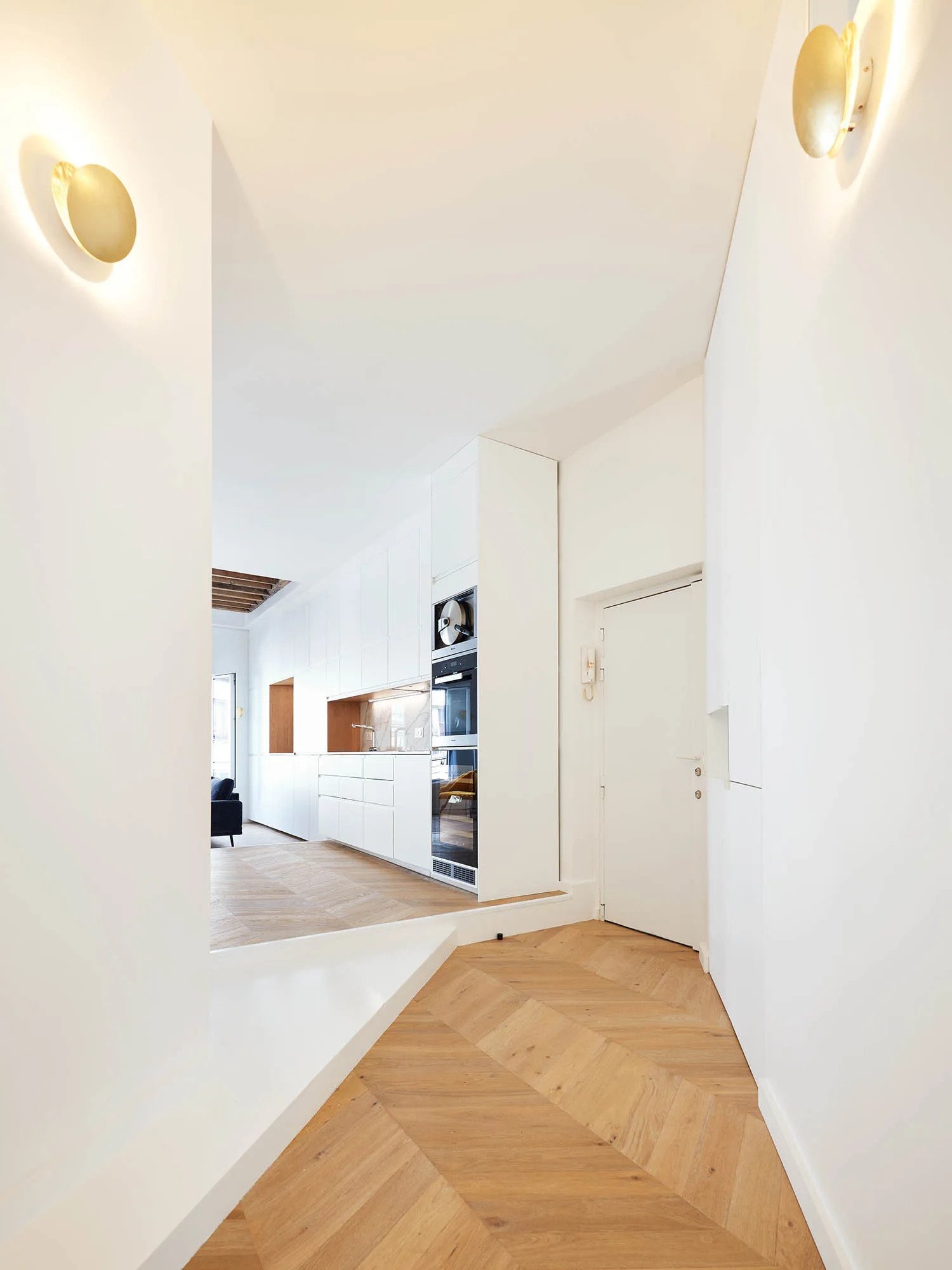 A bright hallway with chevron wood floors leads into a white minimalist kitchen featuring integrated cabinets and gold circular wall sconces.