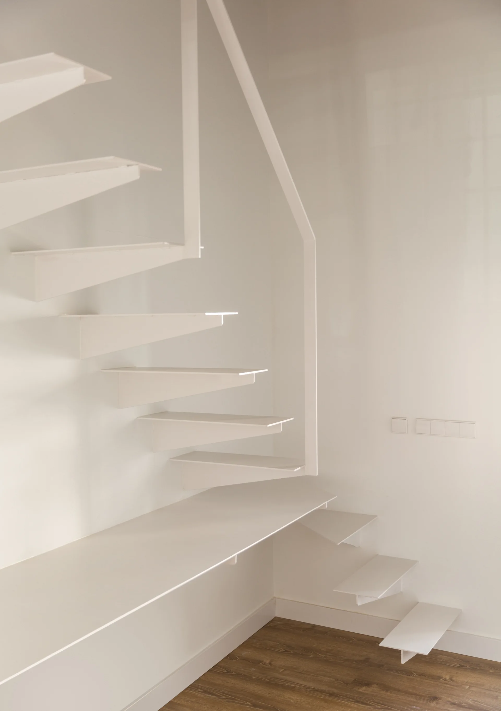 Minimalist white metal floating stairs with an integrated bench rise against a white wall next to wood flooring.