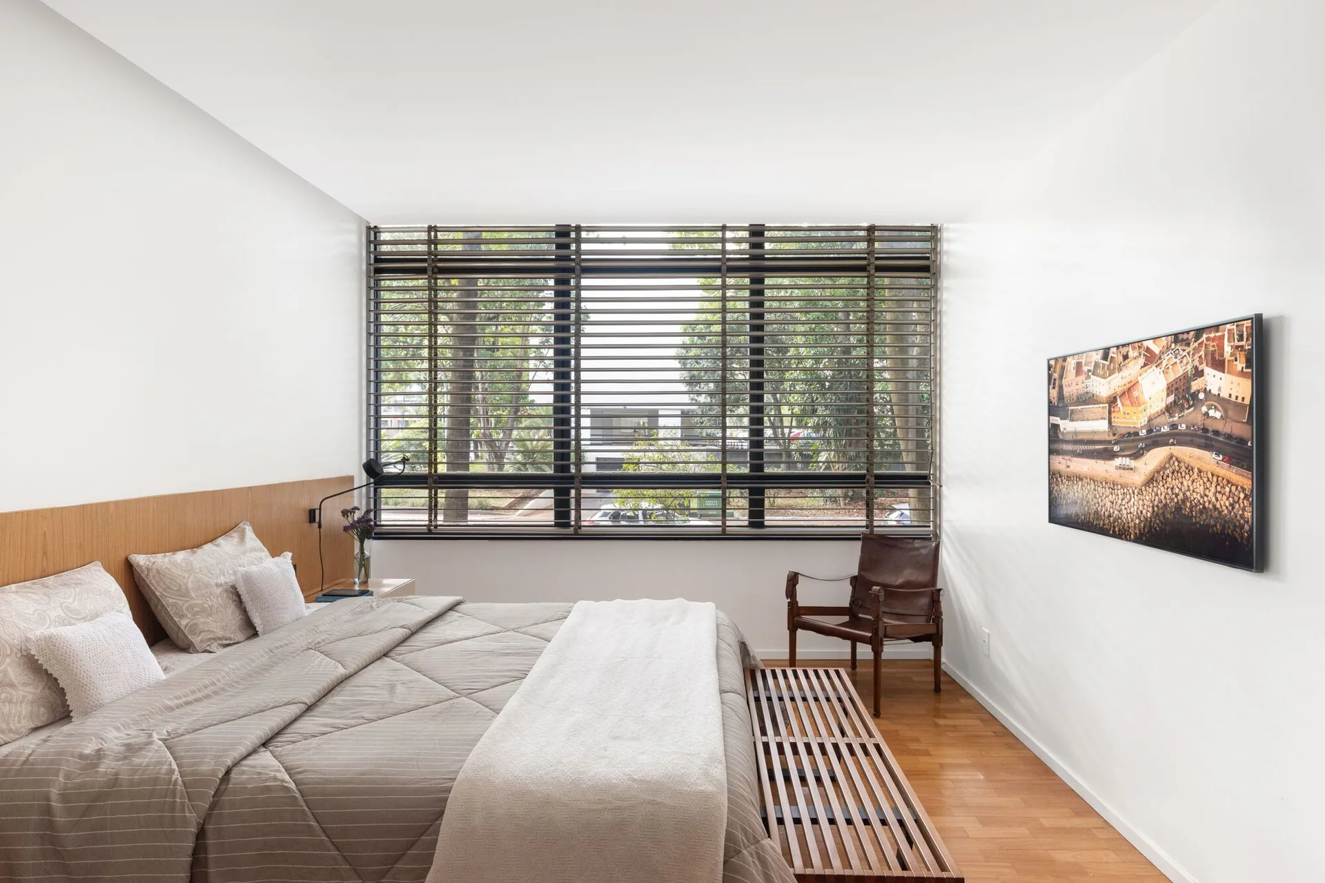 A modern bedroom features a bed with beige bedding, a wooden headboard, a slatted bench, a brown leather armchair, and a large window with blinds.