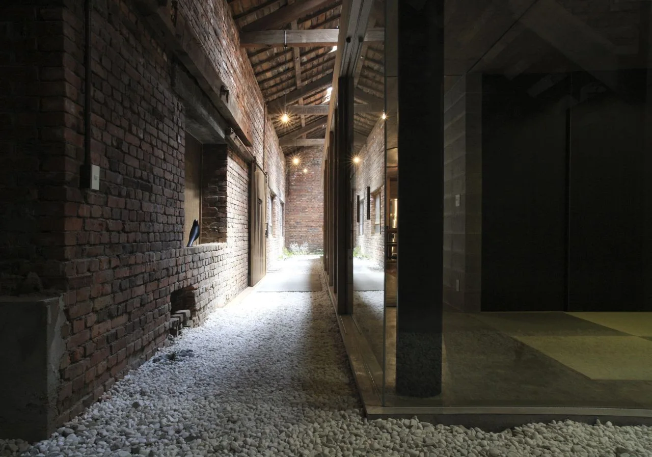 An industrial corridor features a raw brick wall on the left, a white pebble floor, and exposed timber roof beams overhead.