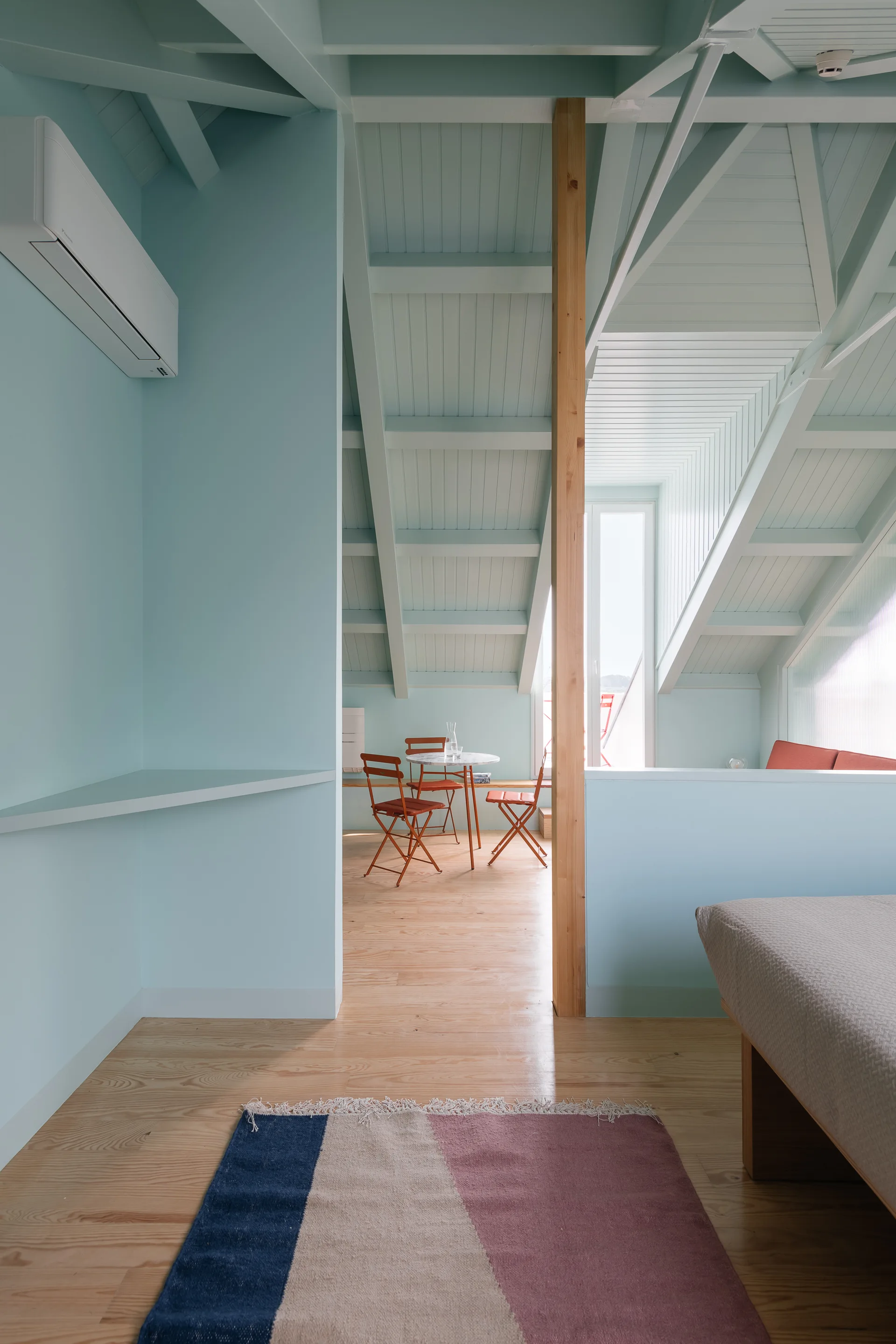 A light blue painted attic loft features vaulted ceilings with exposed beams, light wood floors, and a small dining setup with terracotta chairs.