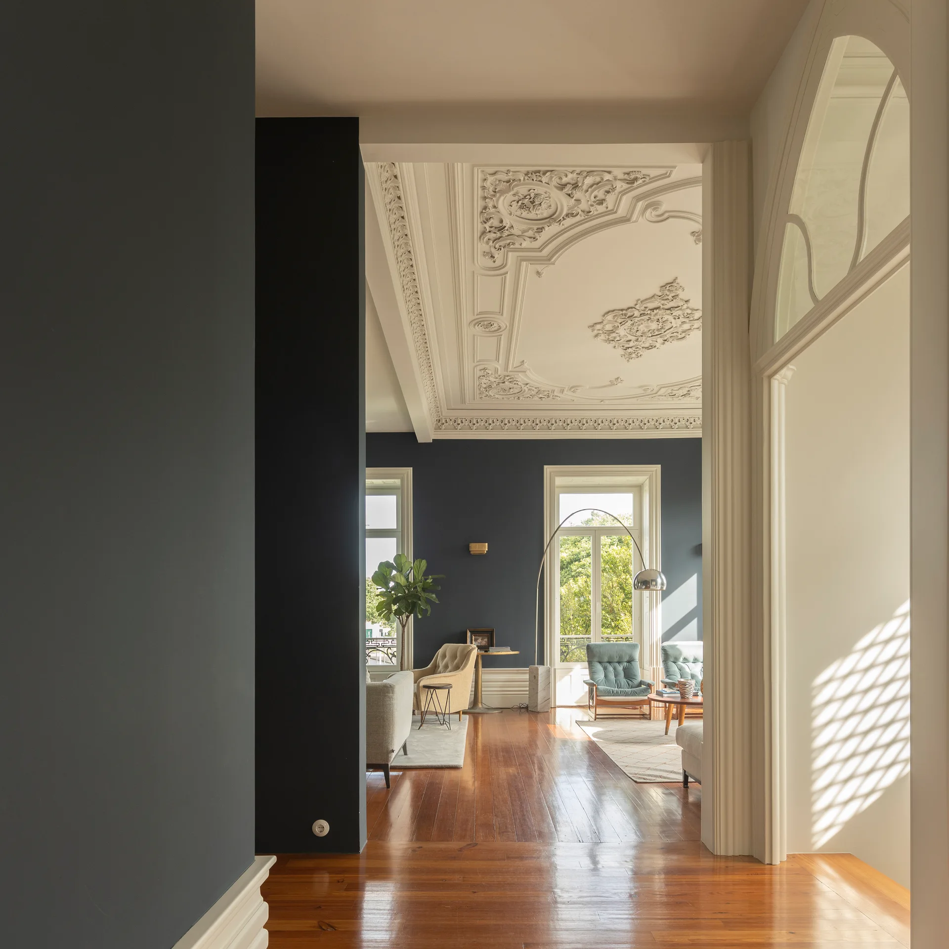 An entryway view looking into a living room with deep charcoal walls, a highly detailed white plaster ceiling, and polished wood floors.