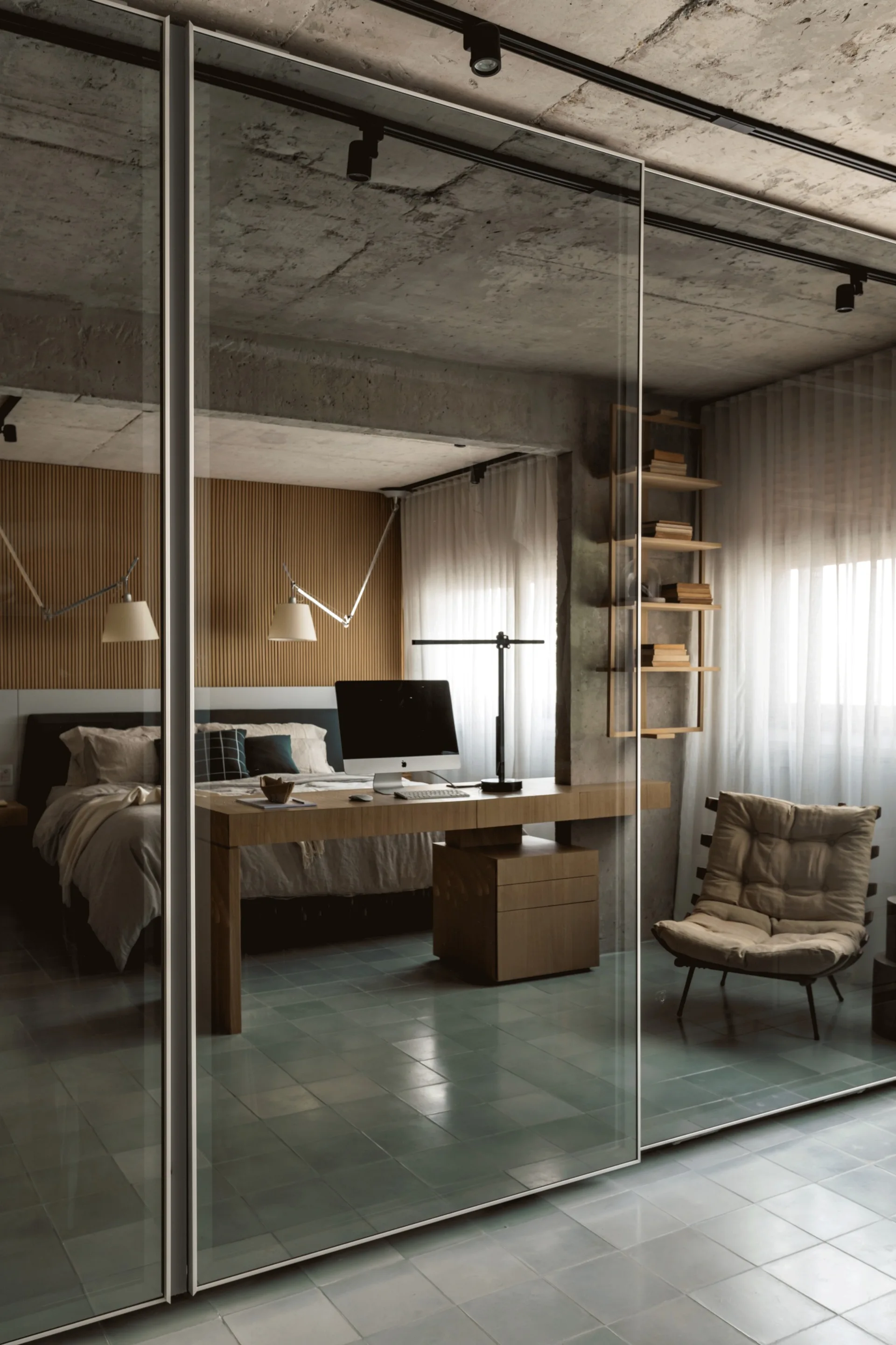 A modern bedroom features a wood desk, a bed with a slatted wood headboard wall, and a large sliding glass partition under an exposed concrete ceiling.