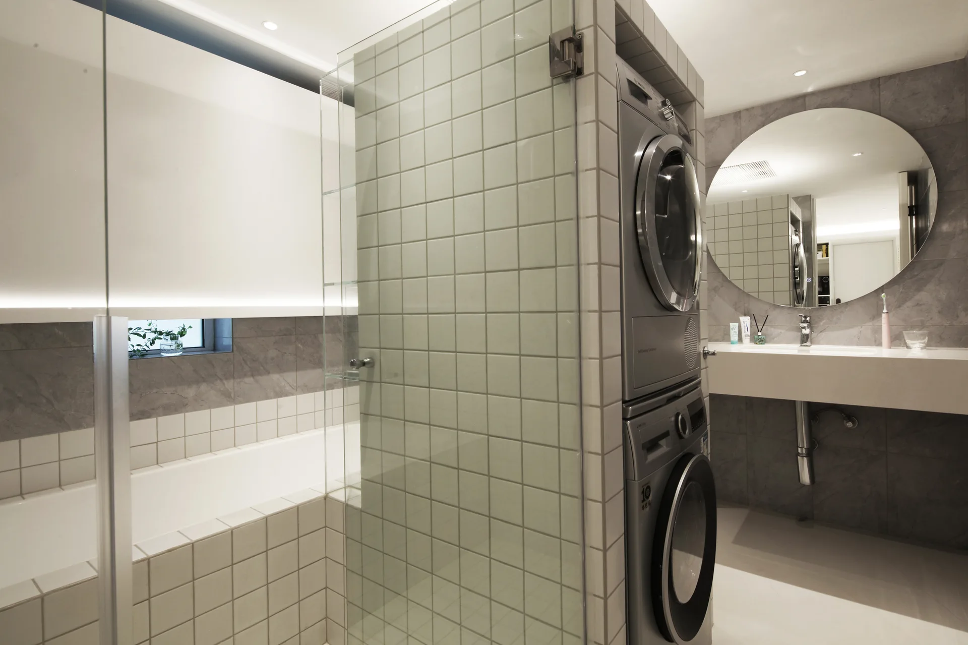 A modern tiled bathroom featuring an integrated stacked washer and dryer next to a glass shower panel and a floating vanity with a round mirror.