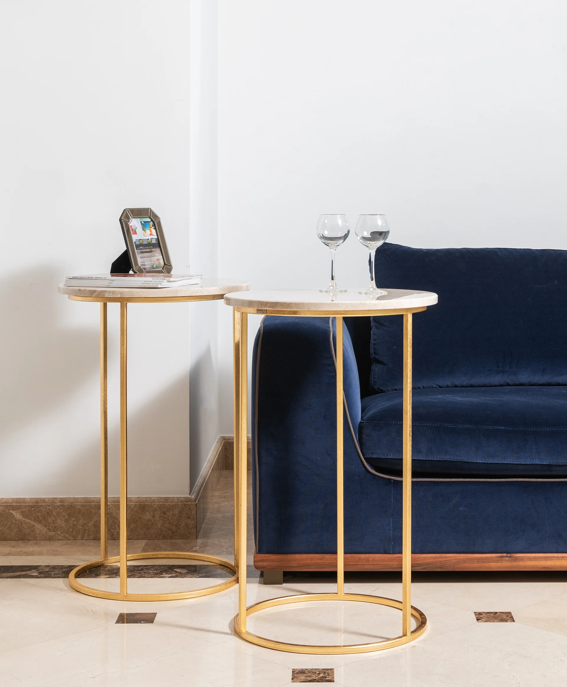 Two round gold side tables with white marble tops stand next to a navy blue velvet sofa against a white wall.