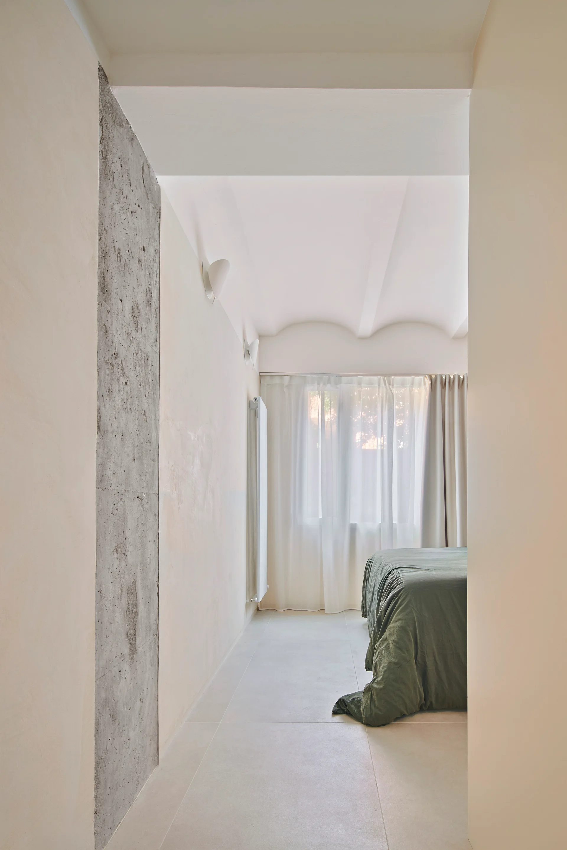 A minimalist bedroom viewed through a doorway, showing an exposed concrete column, soft olive green bedding, and sheer curtains under a vaulted ceiling.