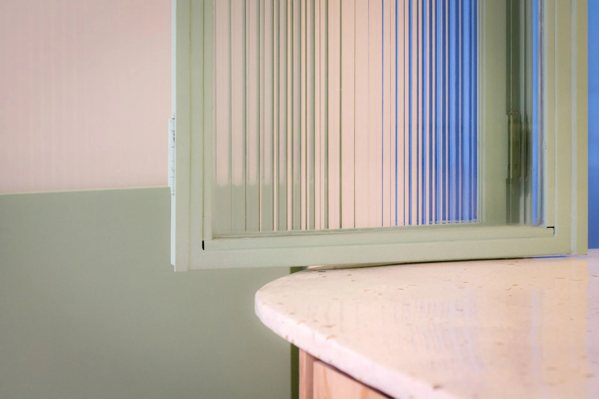 A close-up of an interior window with fluted glass and a sage green frame above a speckled stone countertop and wainscoting.
