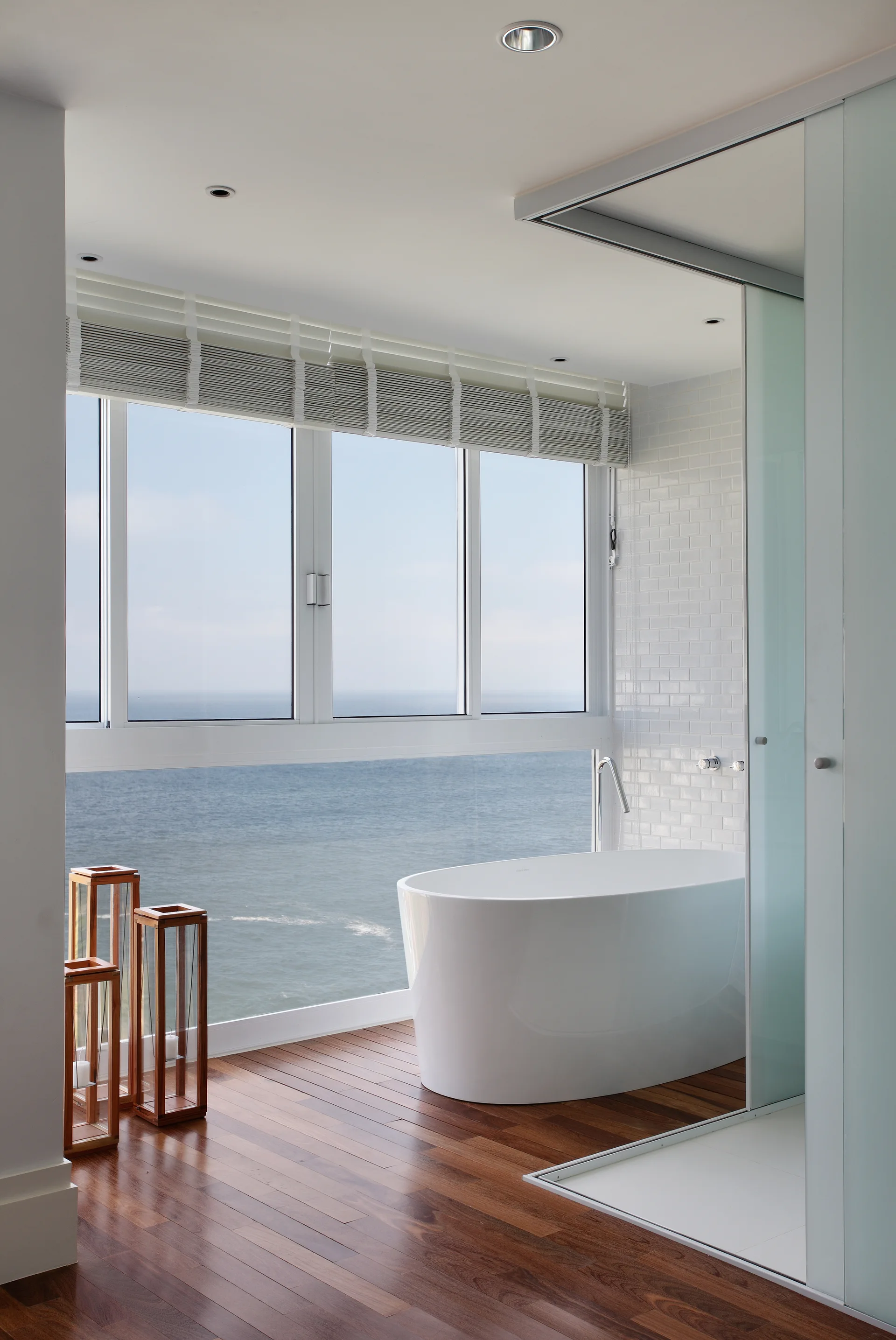 A white freestanding bathtub sits on dark wood floors next to a large window overlooking the ocean in a minimalist bathroom.