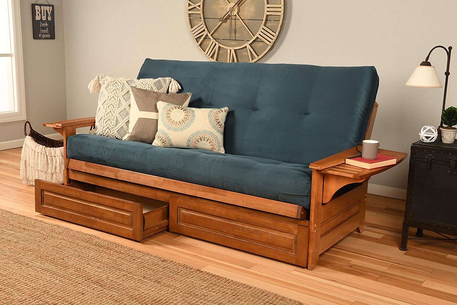 A blue upholstered wooden futon features under-bed storage drawers and a built-in side shelf, positioned in front of a light gray wall with a large clock.