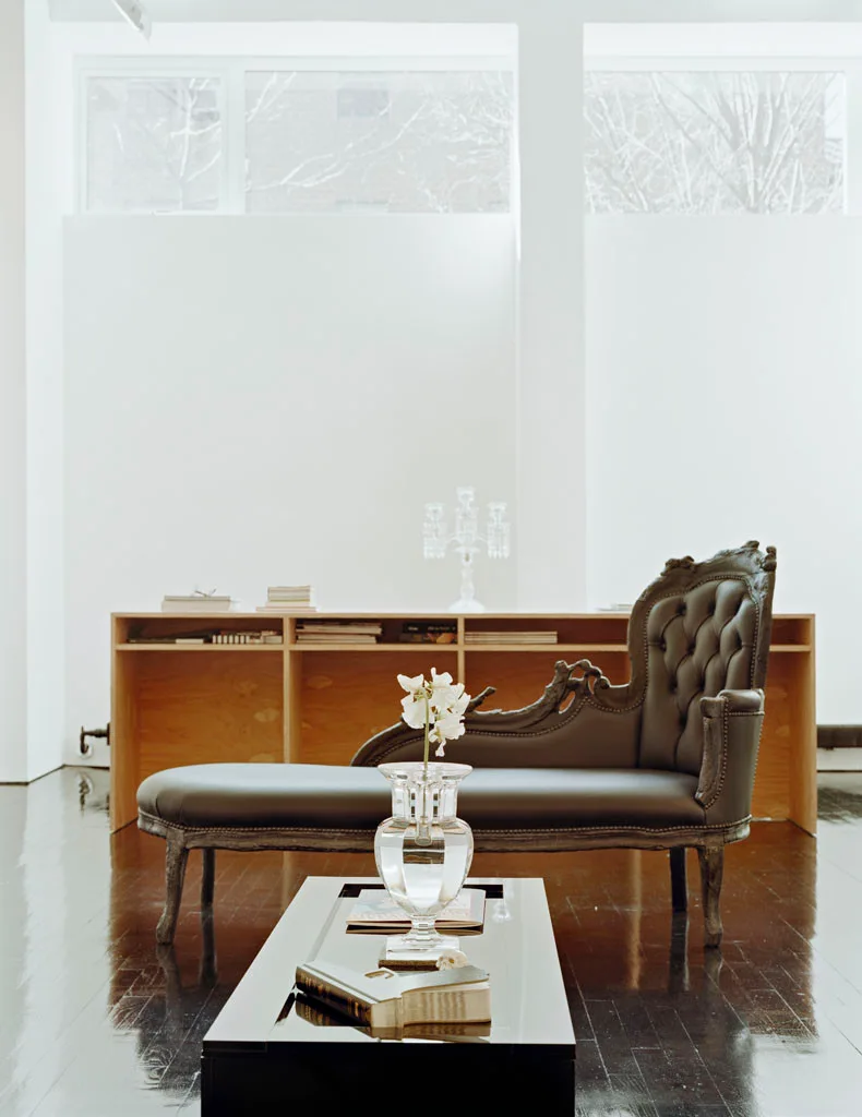 A black tufted chaise lounge sits on dark glossy wood floors in front of a light wood console table and white walls.