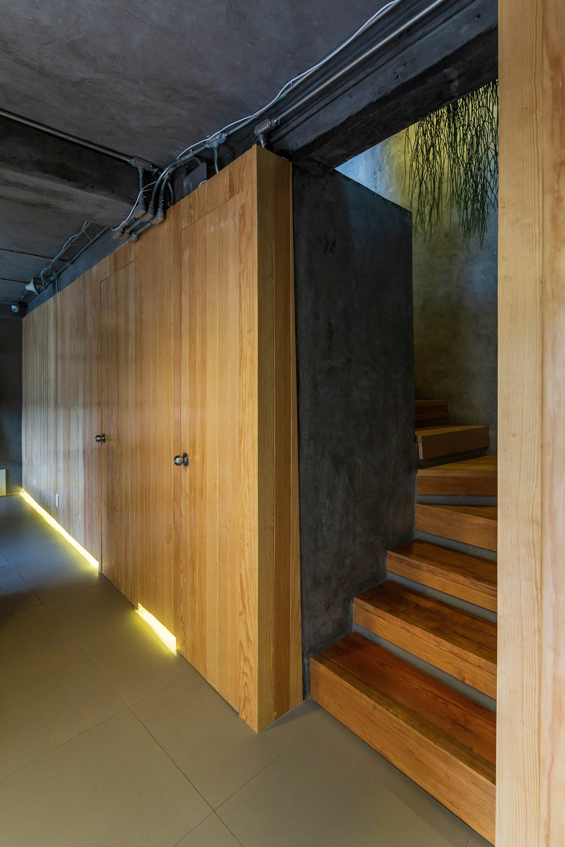 An industrial hallway features wood-paneled storage doors, a concrete ceiling with exposed conduits, and a wooden staircase illuminated by warm floor lighting.