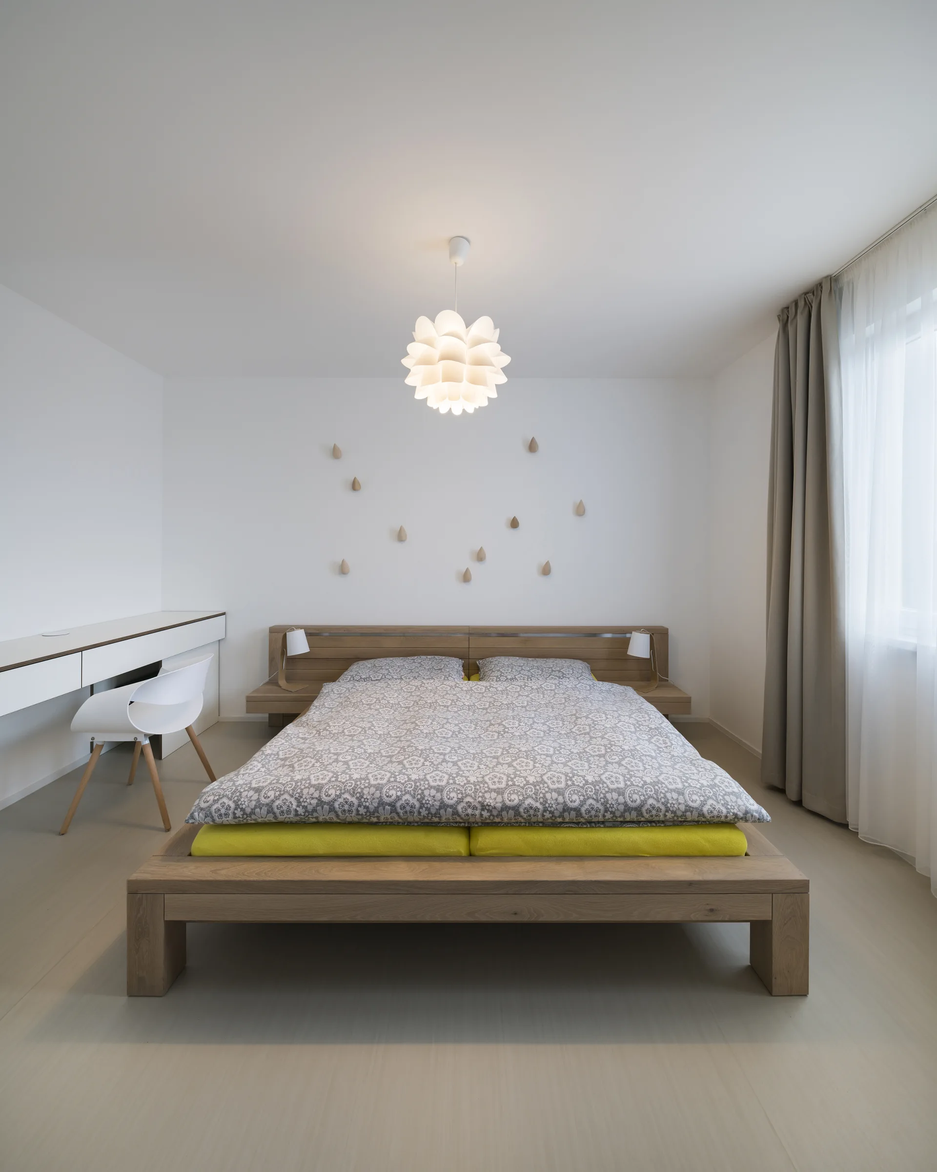A minimalist bedroom featuring a low wooden platform bed, a white floating desk with a modern chair, and small teardrop wall decor.