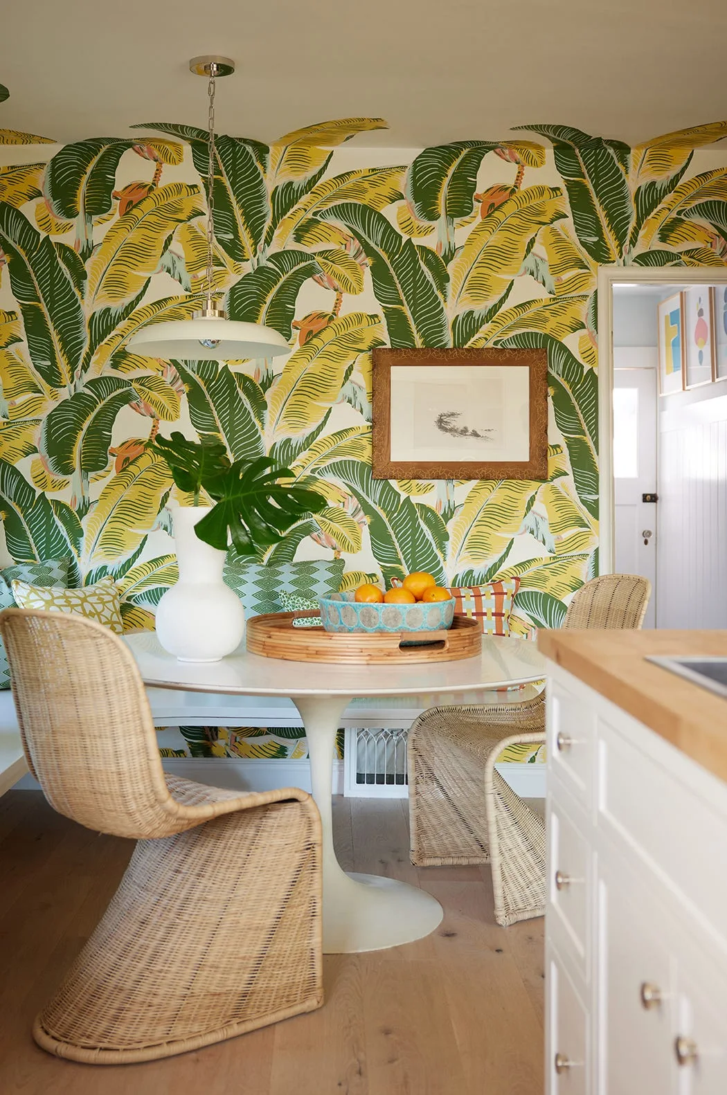 A dining nook features bold banana leaf wallpaper behind a round white pedestal table with woven rattan cantilever chairs and a built-in banquette.