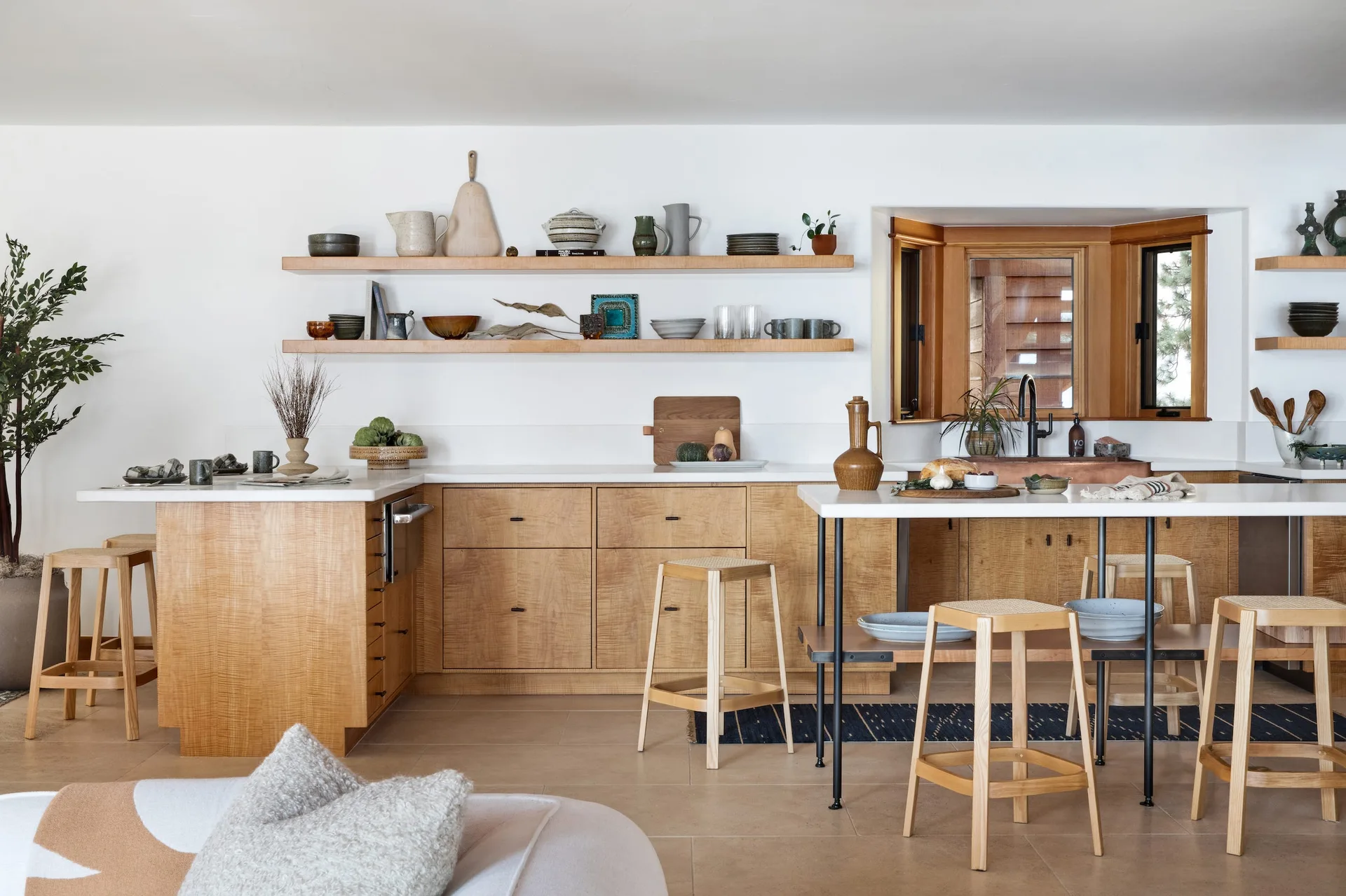 An organic modern kitchen features light wood cabinetry, open floating shelves with ceramics, white countertops, and wooden bar stools with cane seats.