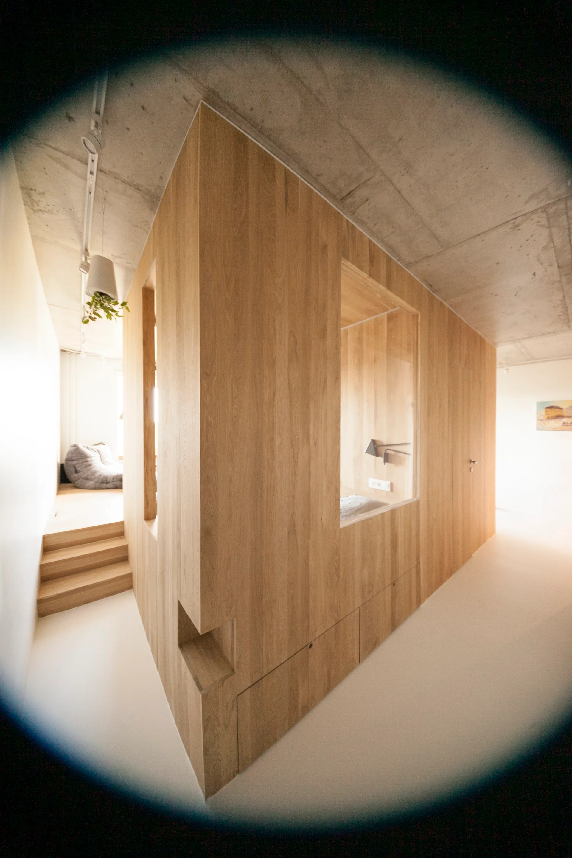 A minimalist wooden partition structure with built-in cutouts sits under an exposed concrete ceiling with a raised platform and steps.