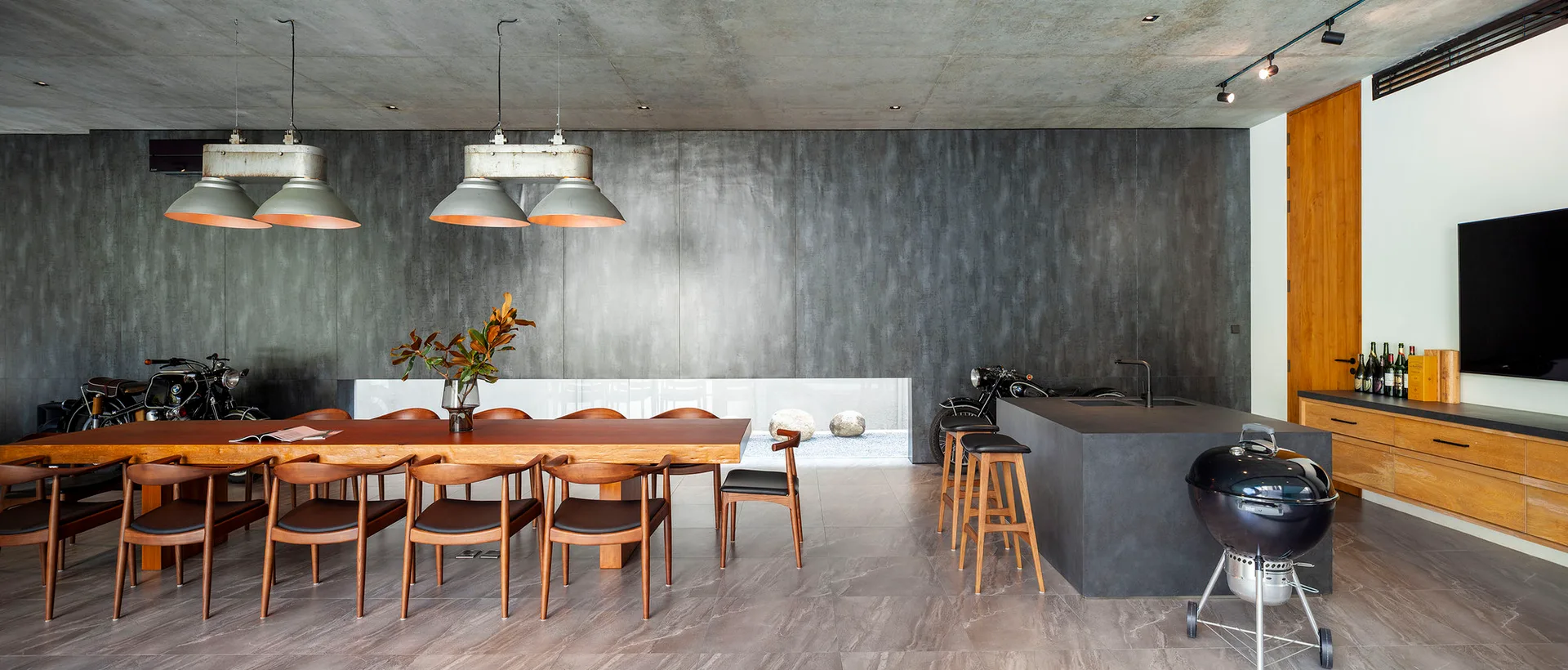 An industrial dining space features a long wooden table with matching chairs, a dark kitchen island, and a raw concrete ceiling with pendant lights.
