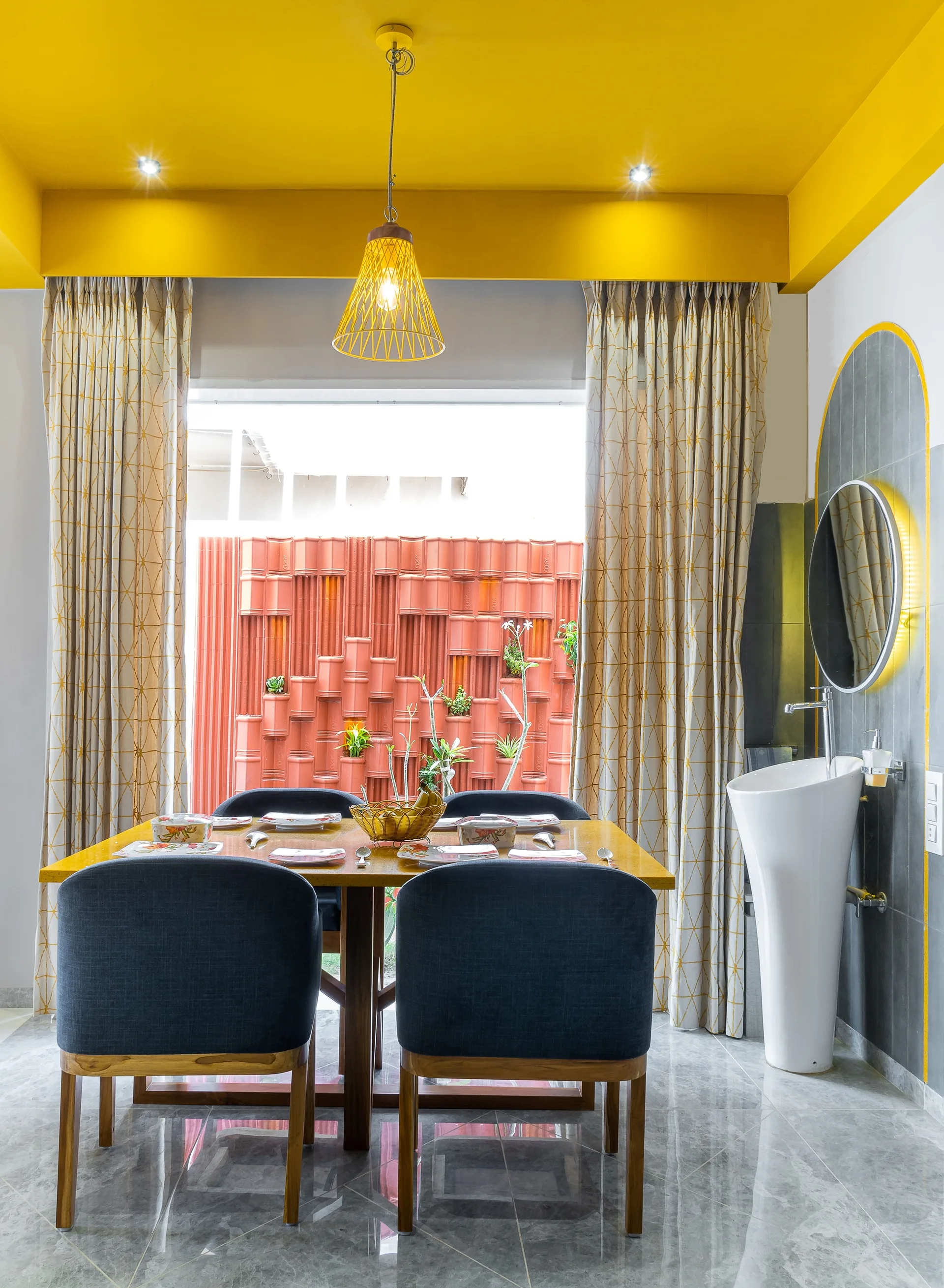 An eclectic dining room features a bright yellow ceiling, a wooden dining table with blue chairs, and a white pedestal sink against a gray tiled wall.