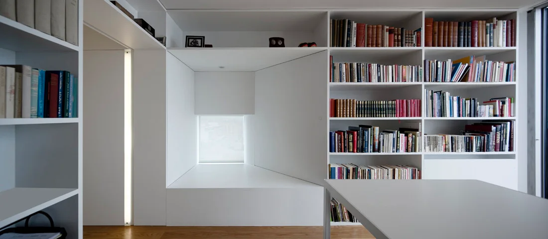 A minimalist white library features built-in floor-to-ceiling bookshelves, a recessed reading nook with a square window, and a vertical strip light.