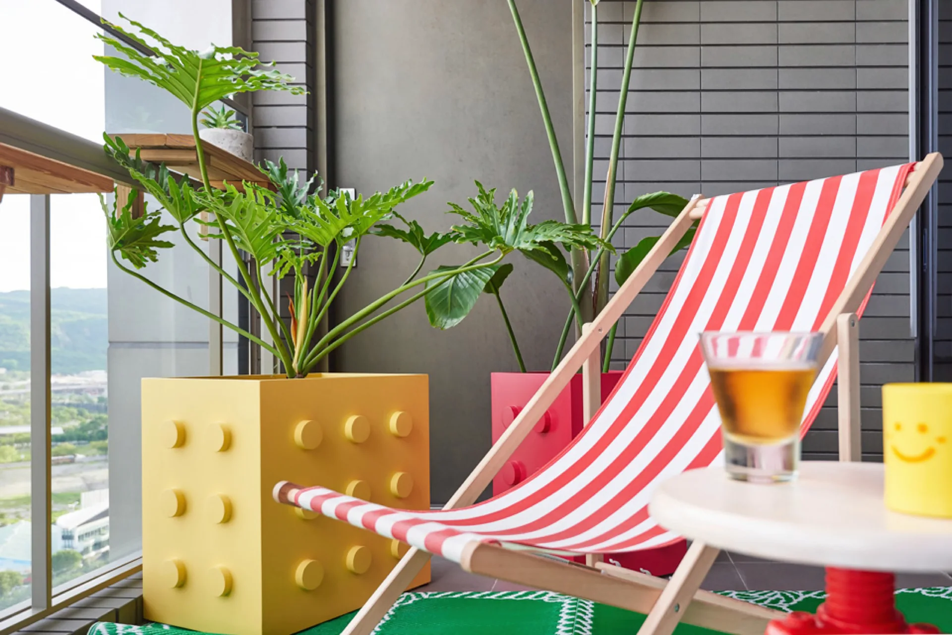 A red and white striped deck chair sits on a balcony next to oversized yellow and red building block planters holding green plants.