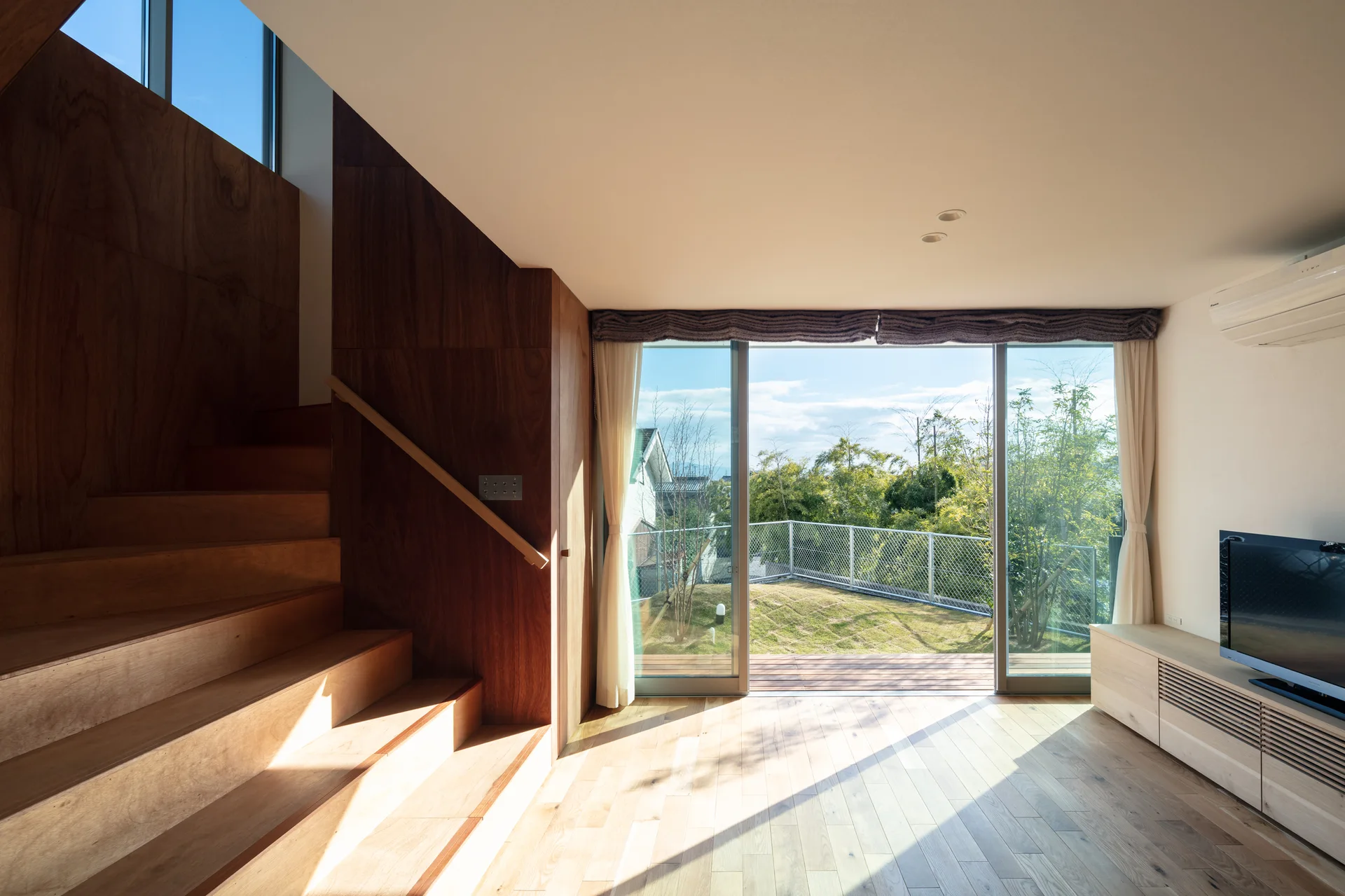 A minimalist living room features a warm wood staircase, light wood floors, and a large sliding glass door opening onto a grassy outdoor garden.
