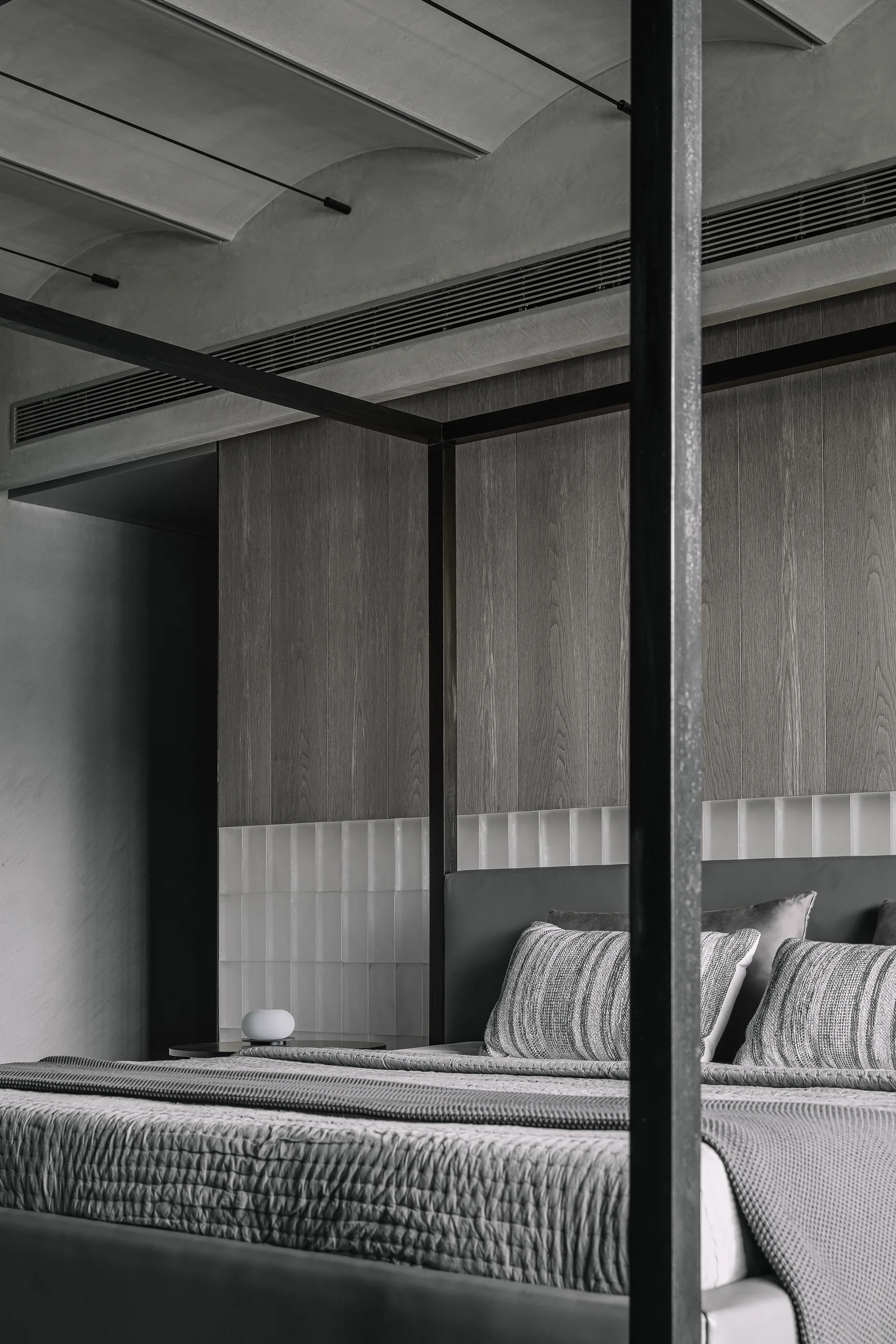 A modern bedroom features a black metal canopy bed set against a vertical wood paneled wall with a row of glossy white tiles.