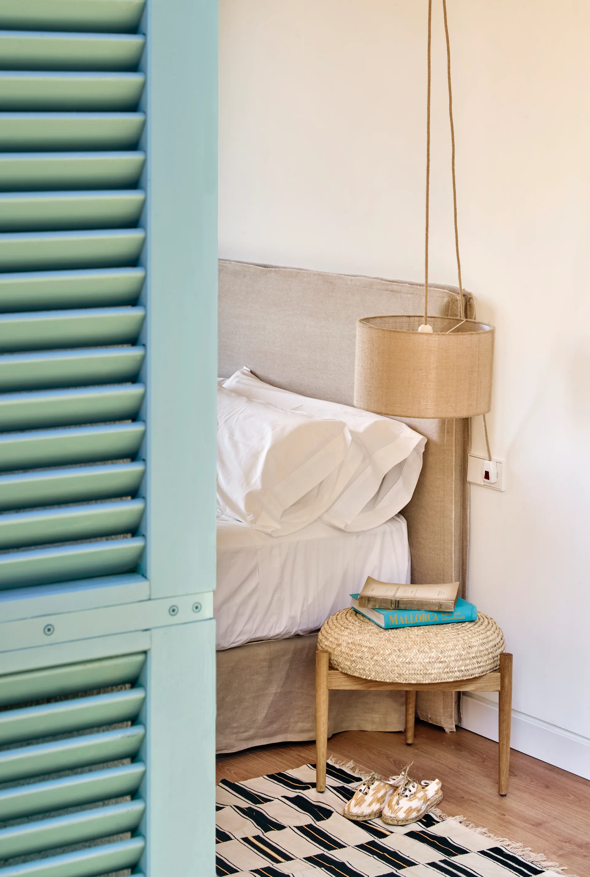 A neutral bedroom corner features a beige headboard, a woven stool nightstand with books, a hanging pendant lamp, and a blue louvered shutter.
