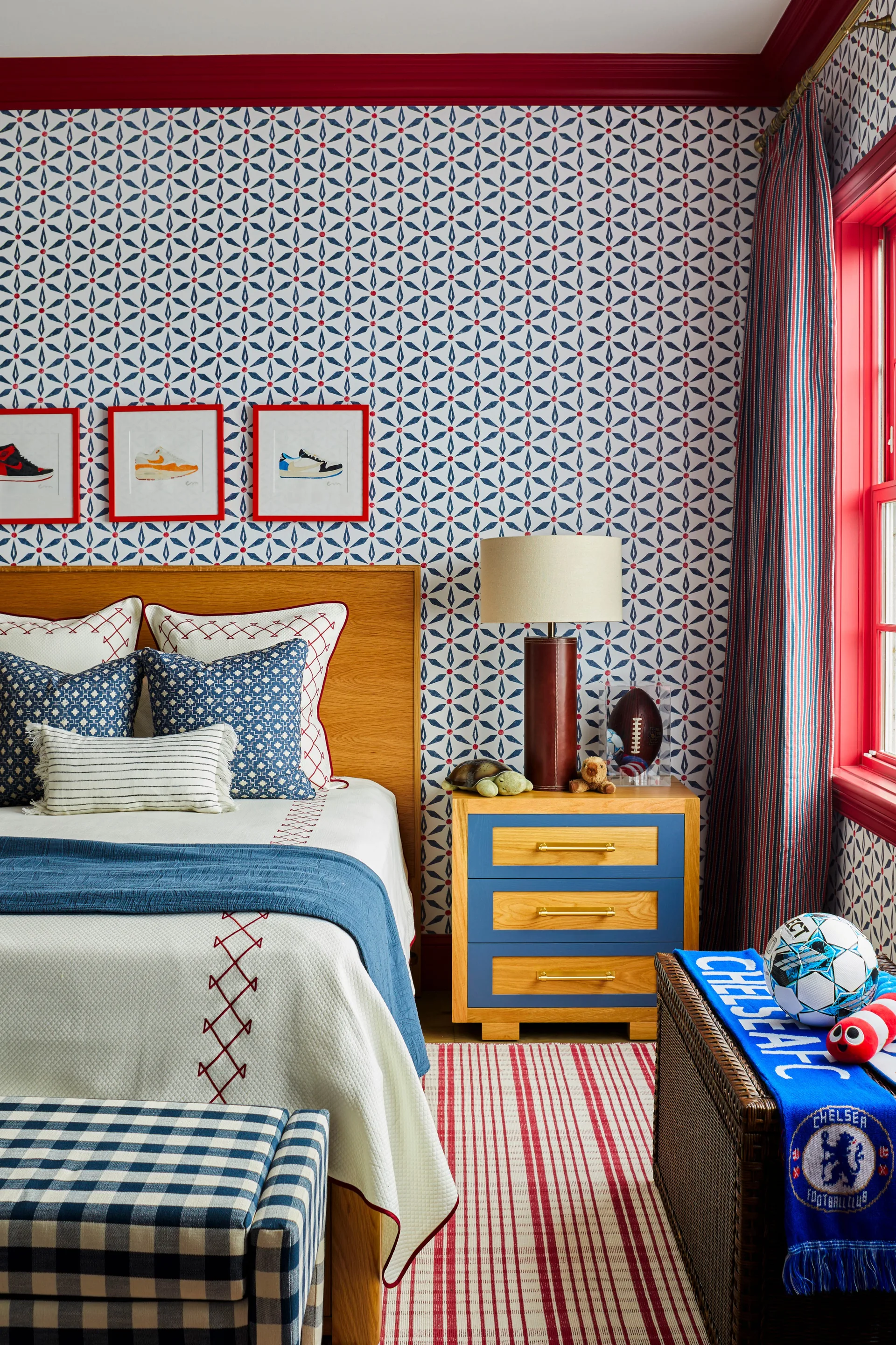 A kids bedroom features geometric wallpaper, a wooden bed, a blue and wood nightstand, and red painted trim around the ceiling and window.