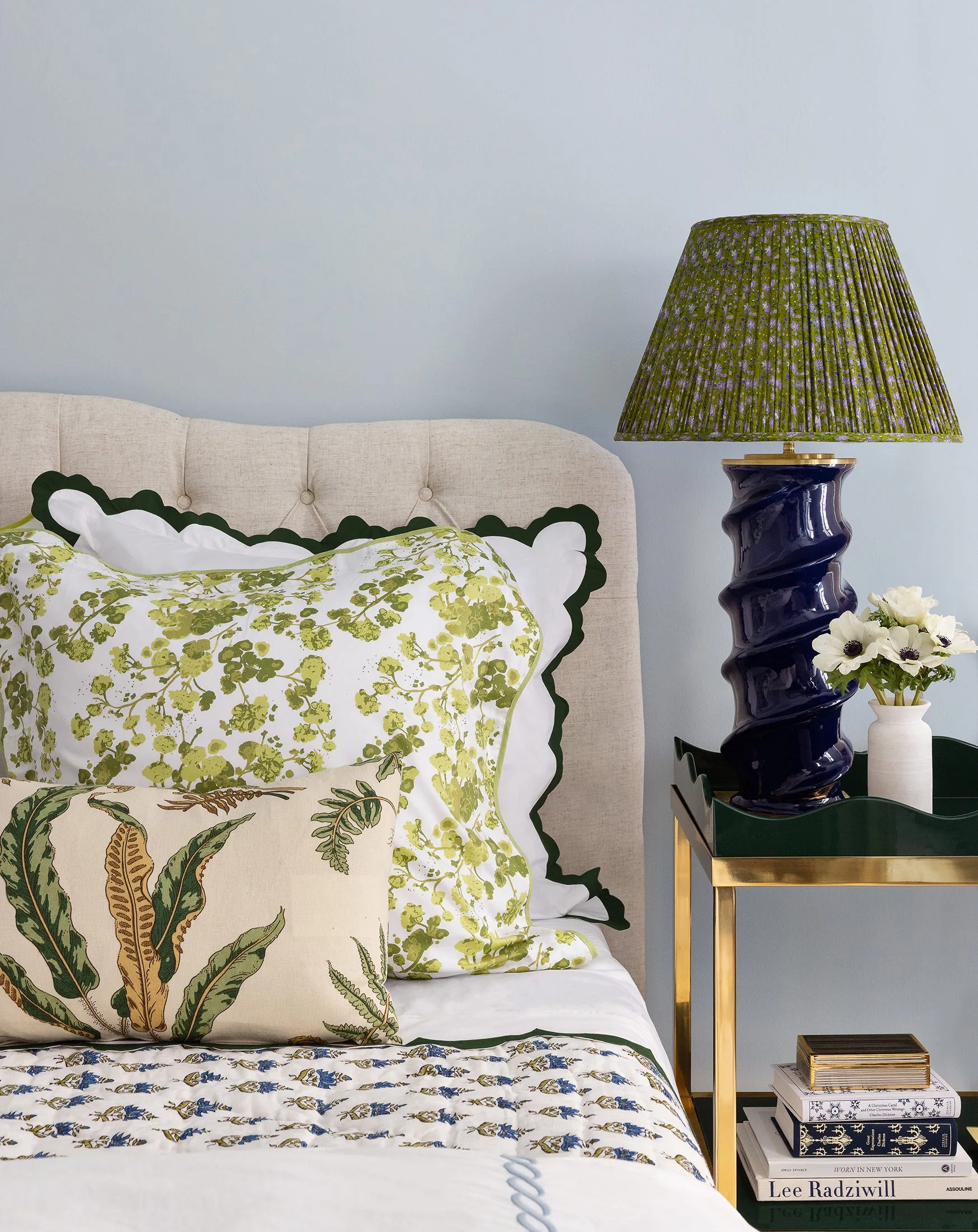 A bedside scene featuring a tufted beige headboard, floral pillows, a dark blue twisted ceramic lamp, and a green scalloped tray table.