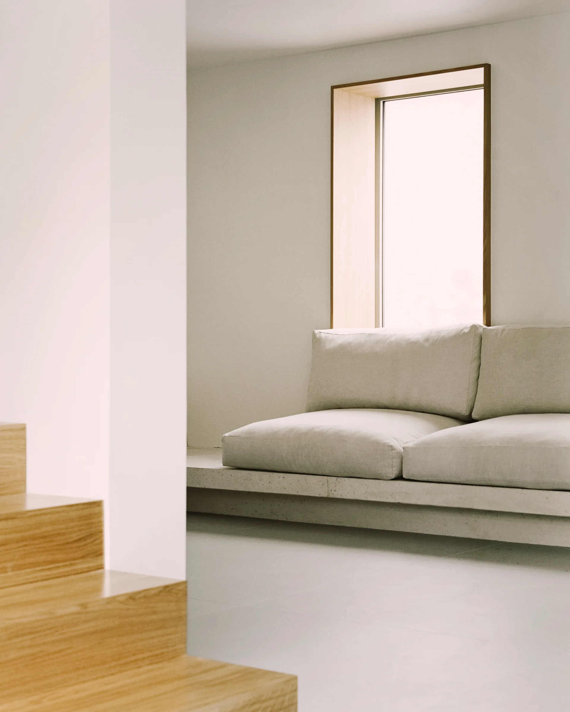 A low-profile beige sofa sits on an integrated concrete platform next to a deep-set wood-framed window, with wooden steps visible in the foreground.