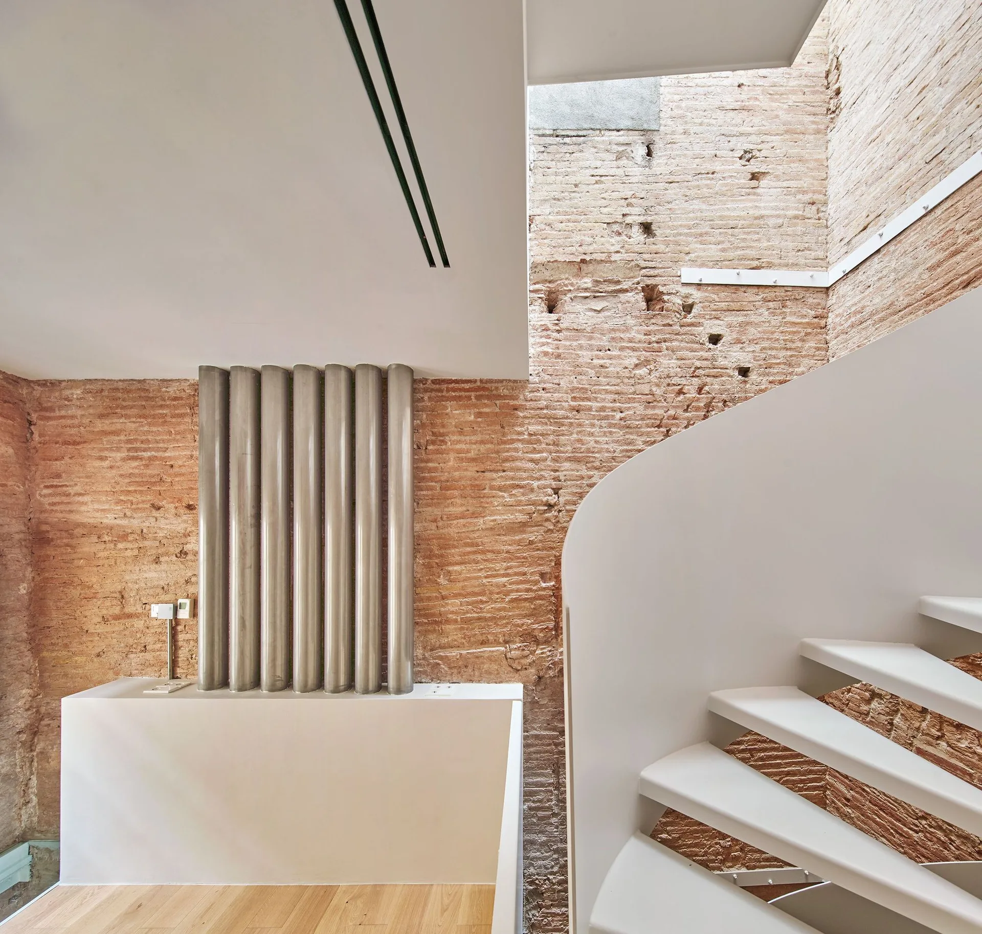 A curved white staircase stands next to a light wood floor and an exposed red brick wall with vertical metallic pipes.
