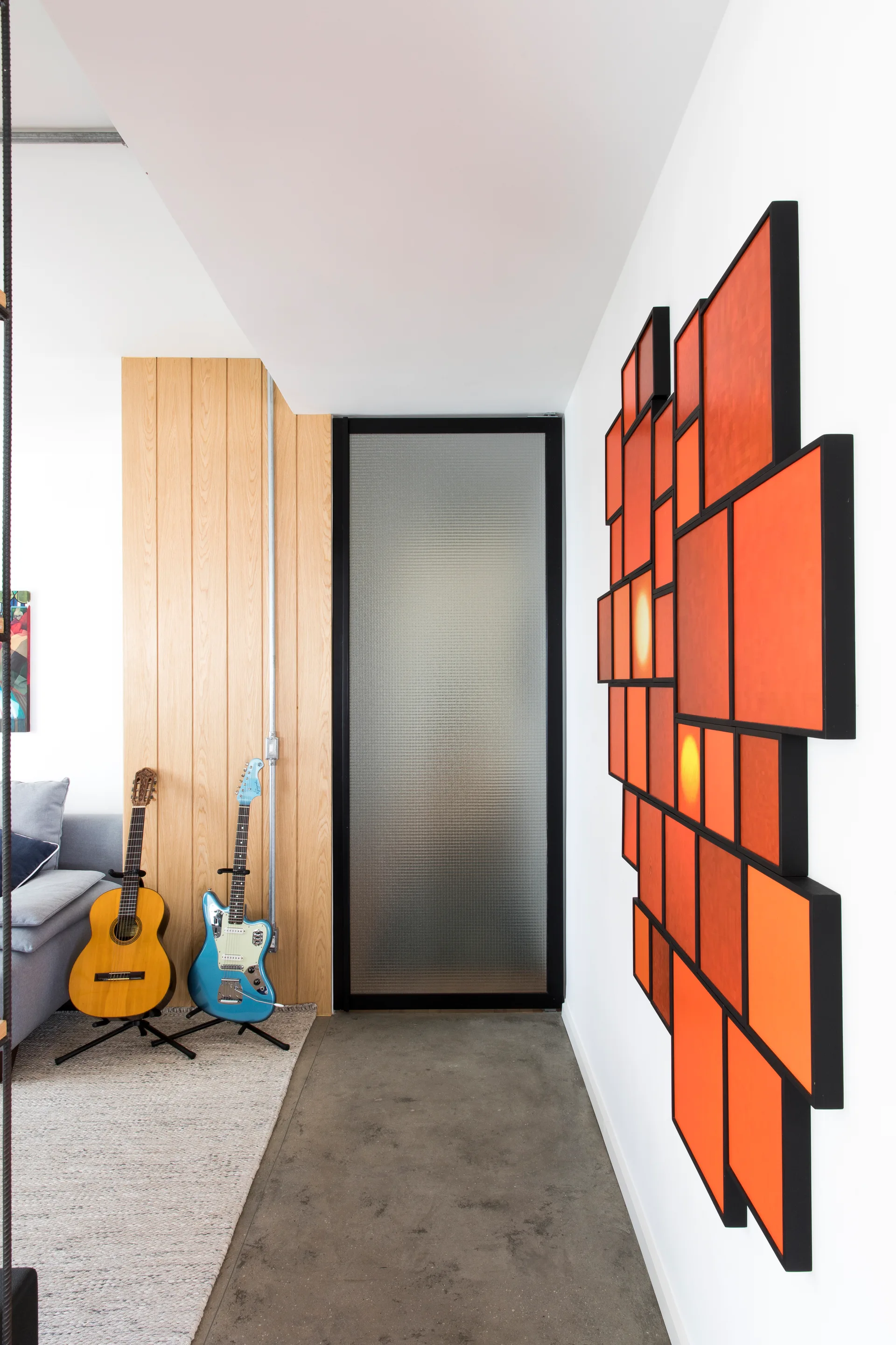 A modern hallway features a textured glass door, warm wood paneling, two guitars on stands, and large orange modular wall art.