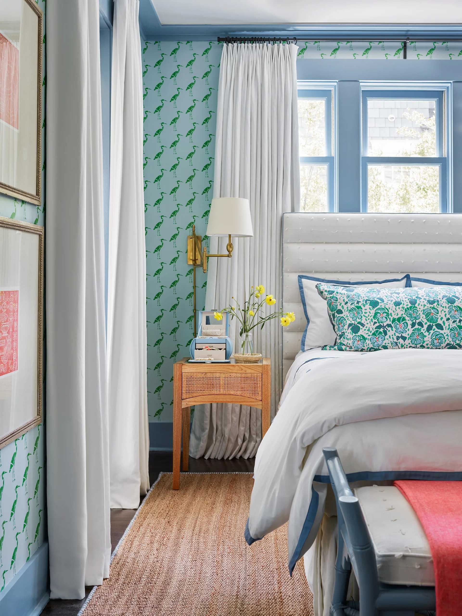 A bedroom featuring light blue bird-print wallpaper, an upholstered white headboard, a wooden cane nightstand, and a brass wall sconce.