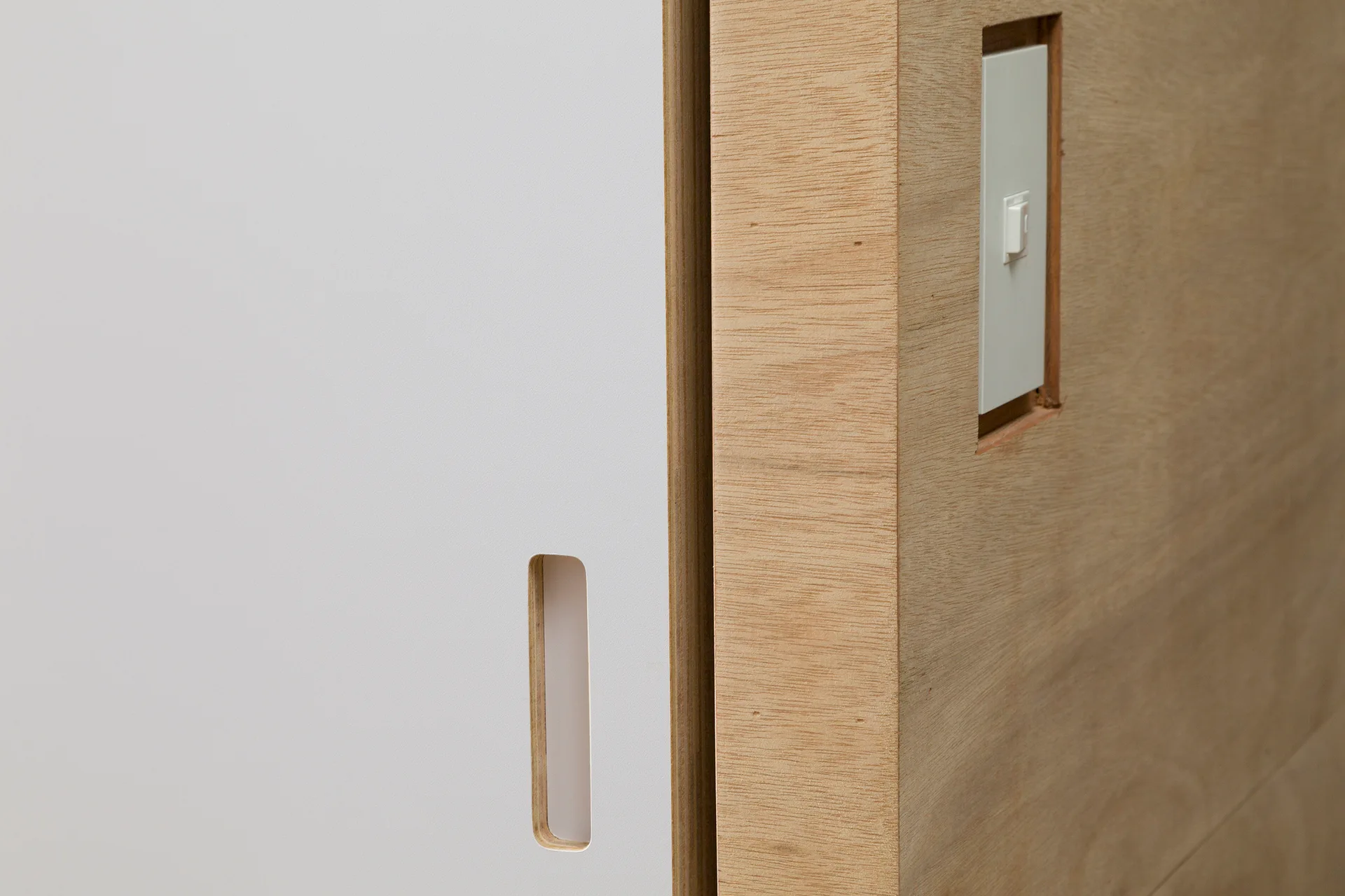 A close-up of a white sliding door with a recessed handle next to a plywood wall with an integrated white light switch.