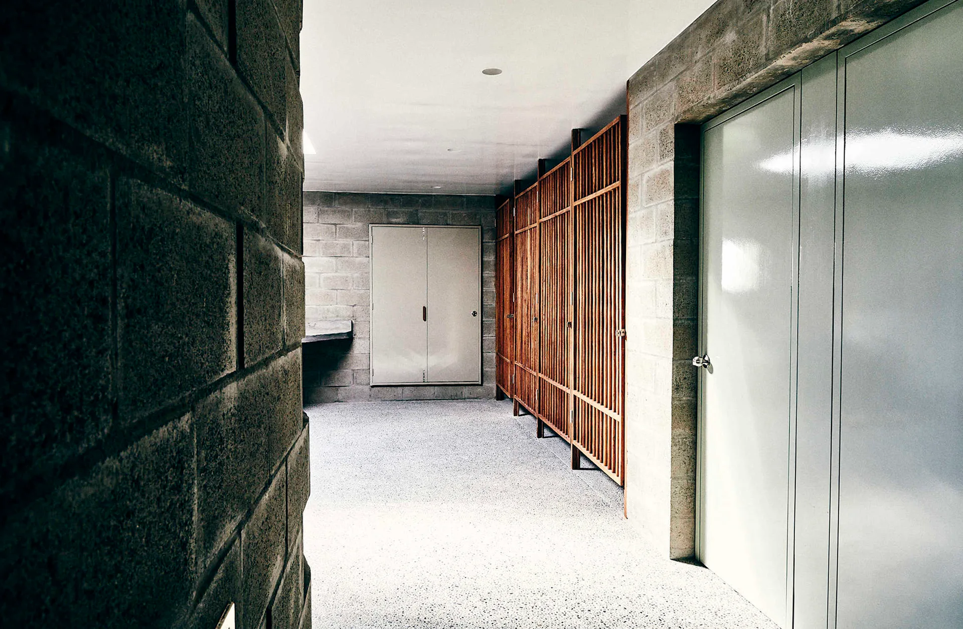 A hallway featuring exposed gray concrete block walls, a light speckled terrazzo floor, and a tall vertical wood slatted screen.