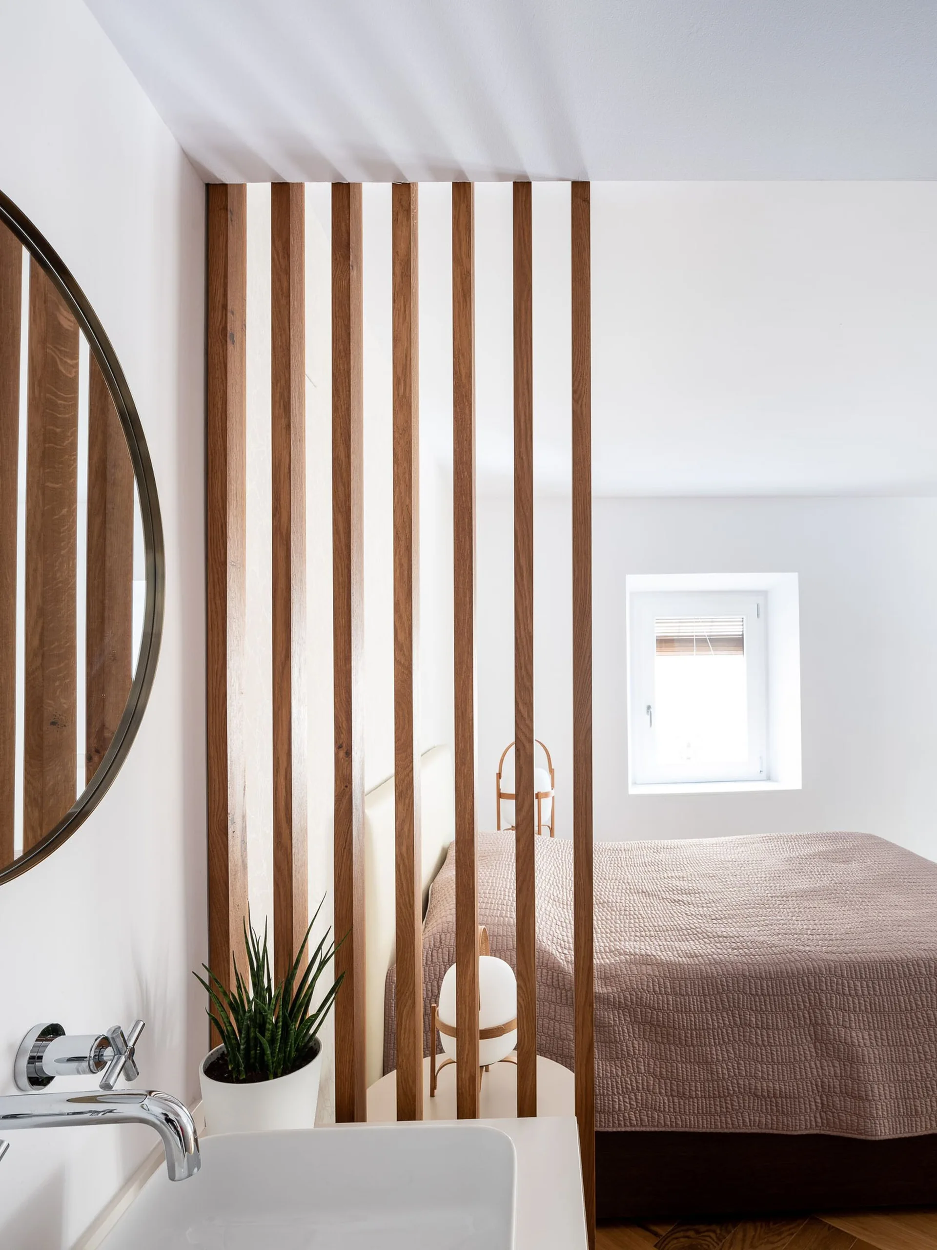 A vertical wood slat partition separates a white bathroom sink in the foreground from a bedroom with a mauve-covered bed in the background.