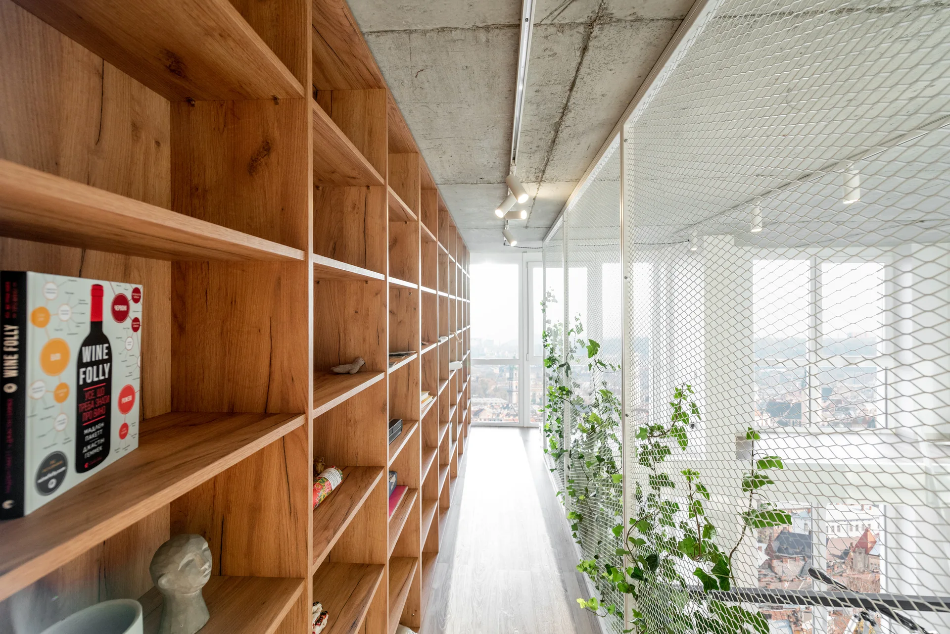 A narrow hallway features a large wood built-in bookshelf on one side and a white metal mesh partition with green climbing plants on the other.