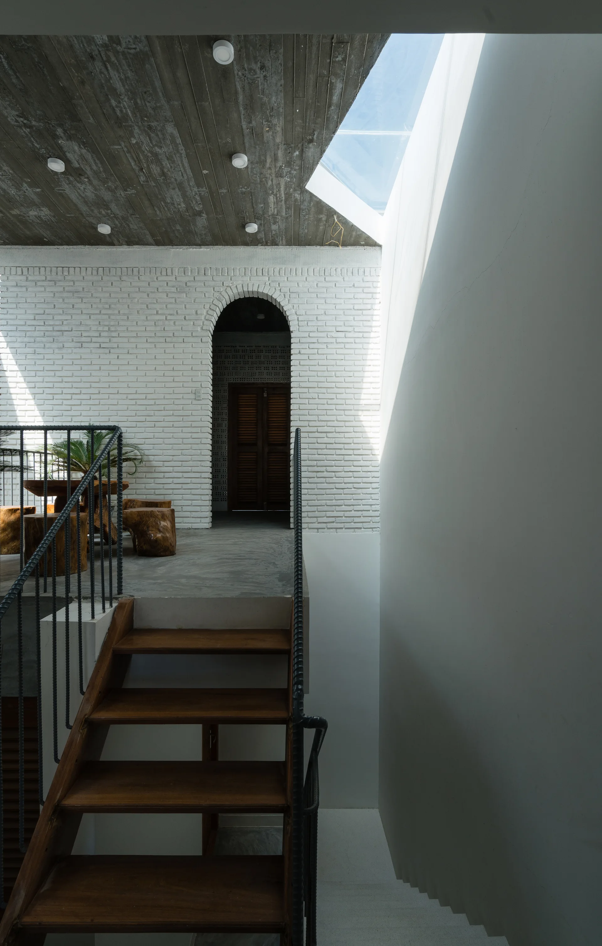 A wooden staircase with black metal railings leads to a concrete landing with a white painted brick wall and an arched doorway under a skylight.