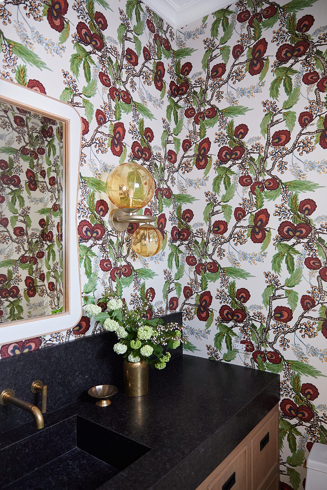 A powder room features a dark stone vanity, a wall-mounted brass faucet, an amber double globe sconce, and bold botanical floral wallpaper.