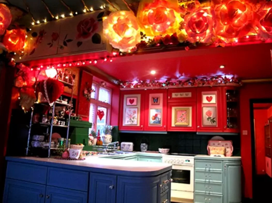 An eclectic kitchen with red upper cabinets, teal lower cabinets, a blue island, and illuminated floral garlands along the ceiling.