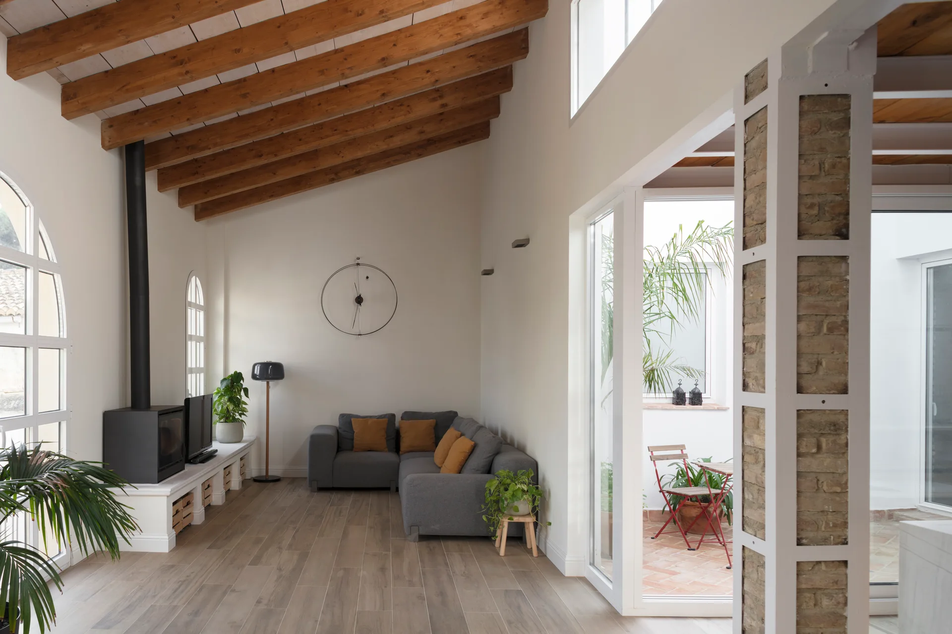 A bright living room features a gray corner sectional, a black fireplace stove on a low console, and exposed wooden ceiling beams.