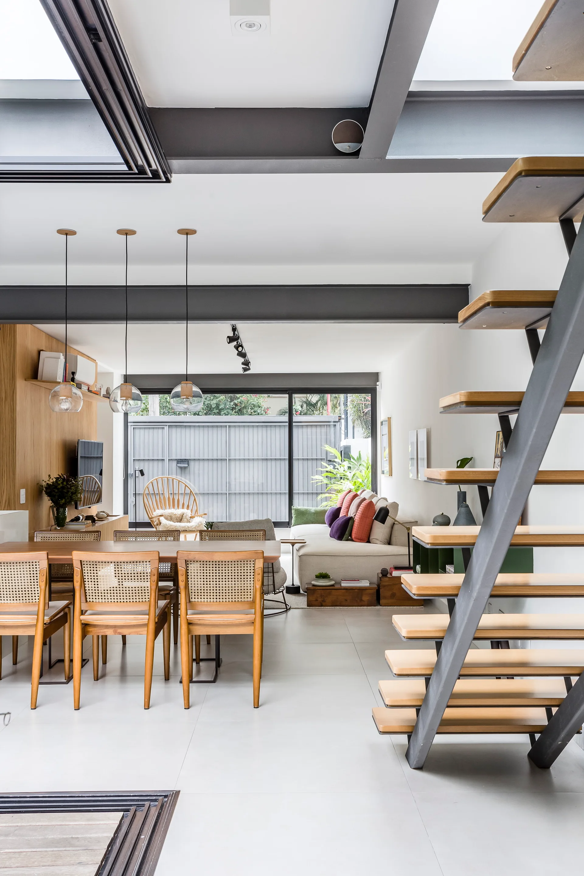 An open-plan interior featuring a wooden dining set with cane chairs, a modern open-riser staircase, and a neutral sofa.