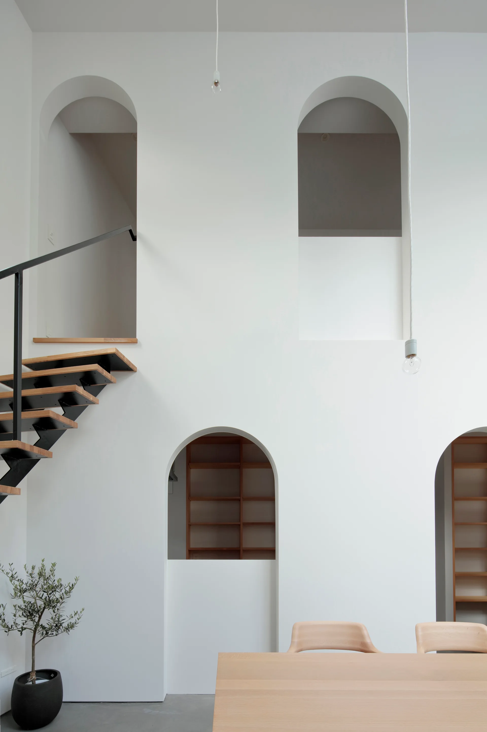 A minimalist interior featuring a white wall with multiple arched openings, a floating wood staircase, and a light wood dining table.