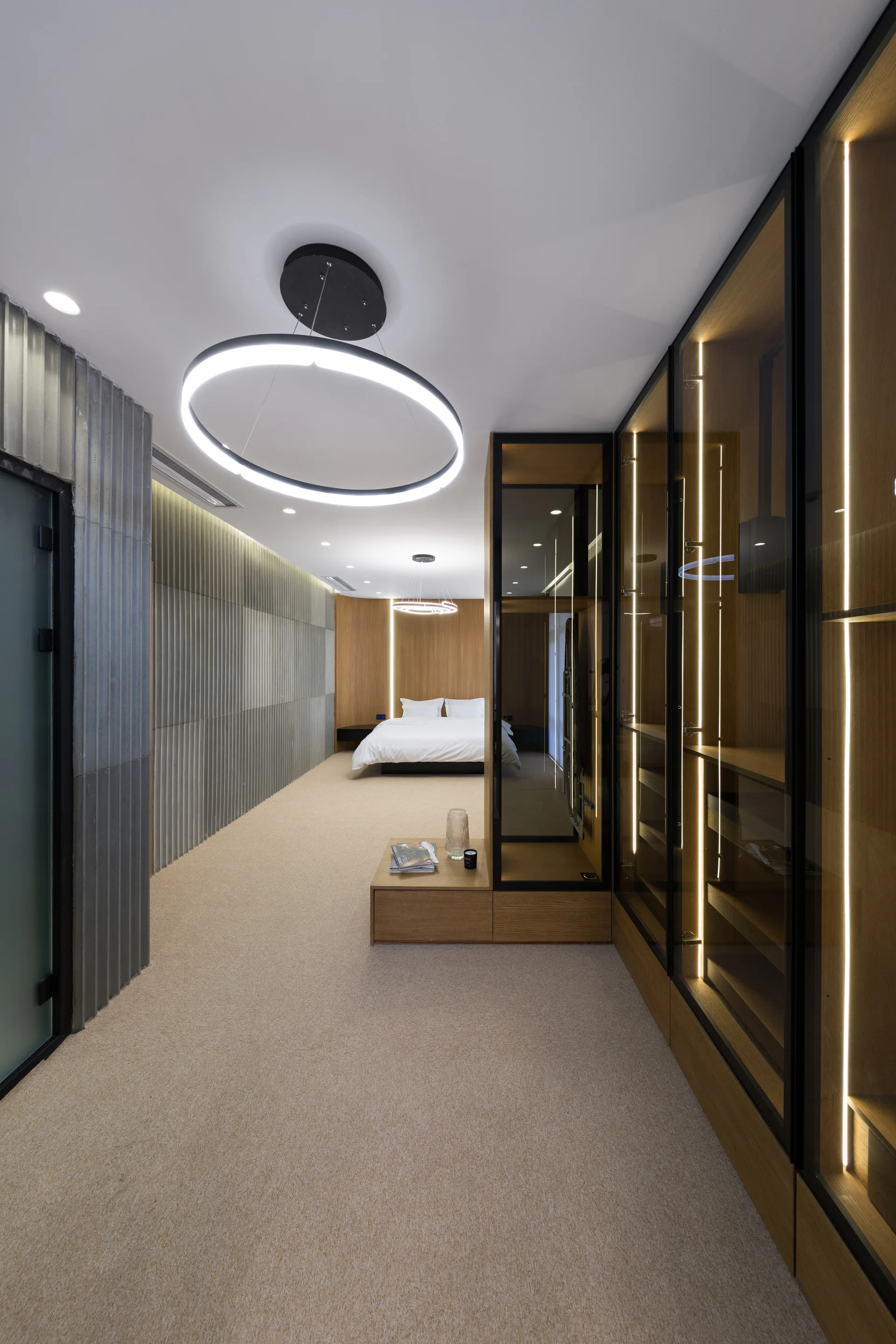 A modern minimalist bedroom featuring a glass-front wardrobe with vertical LED lighting, ribbed metal wall paneling, and circular pendant lights.