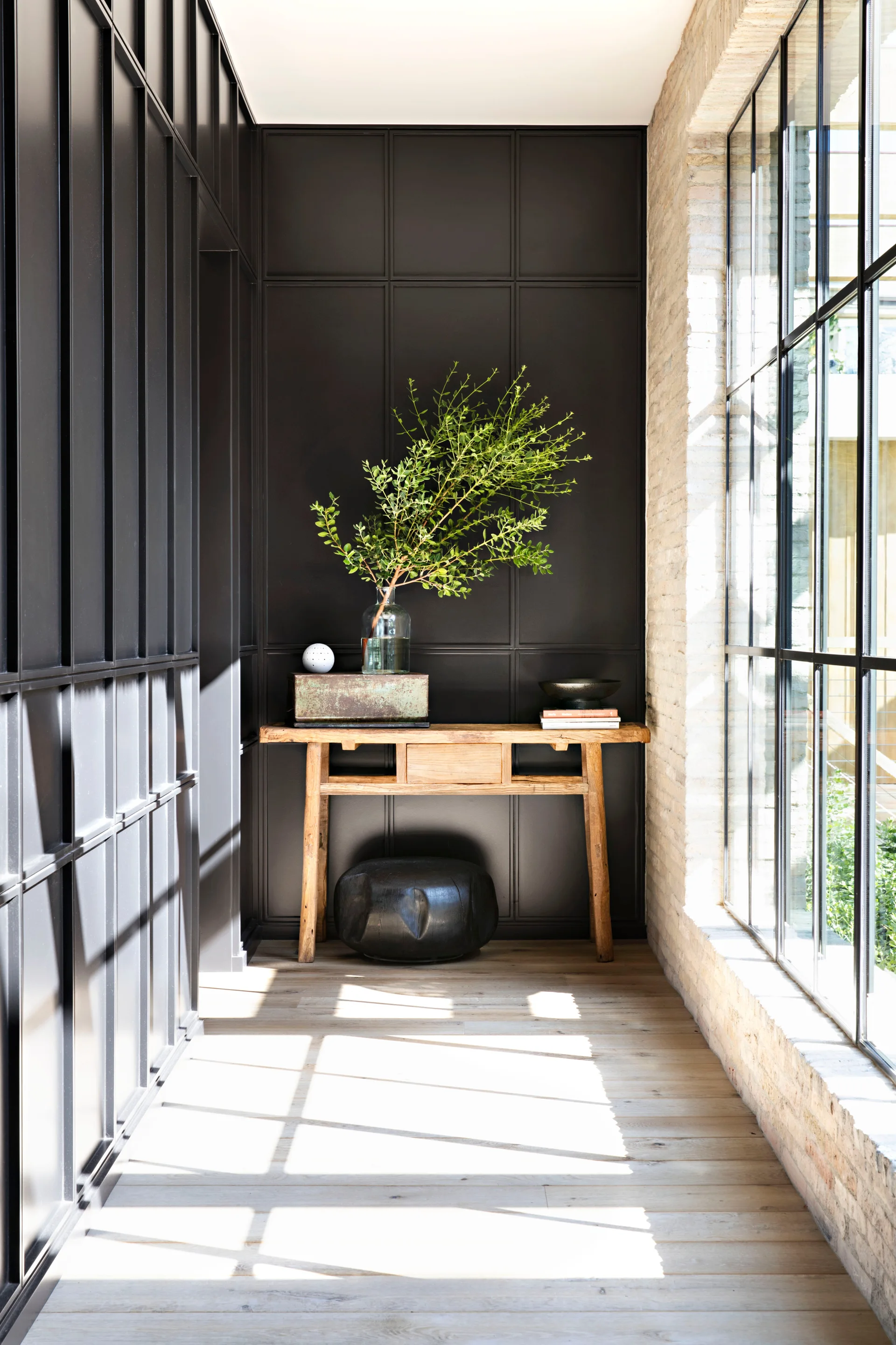 A sunlit hallway features a rustic wooden console table against a matte black paneled wall next to large grid windows set in exposed brick.